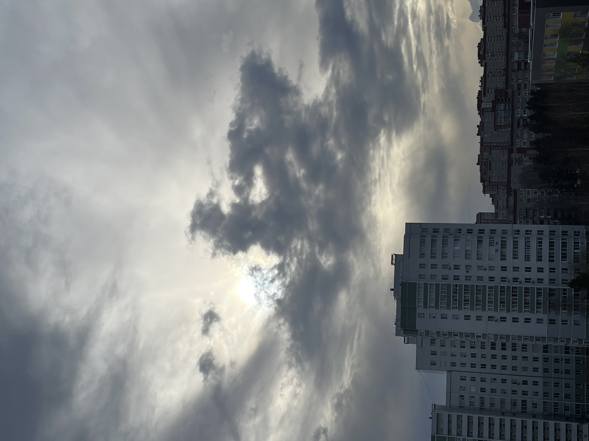 The image depicts a cityscape under an overcast sky. The sun is partially obscured by dark, swirling clouds, creating a diffused light effect. Two tall apartment buildings dominate the right side of the frame. The closest building has a light grey facade with green-trimmed balconies. Further back, a series of older, more dense apartment blocks are visible, appearing in shades of grey and brown. A few trees are interspersed among the buildings. The image appears to be taken in Izhevsk, Russia.