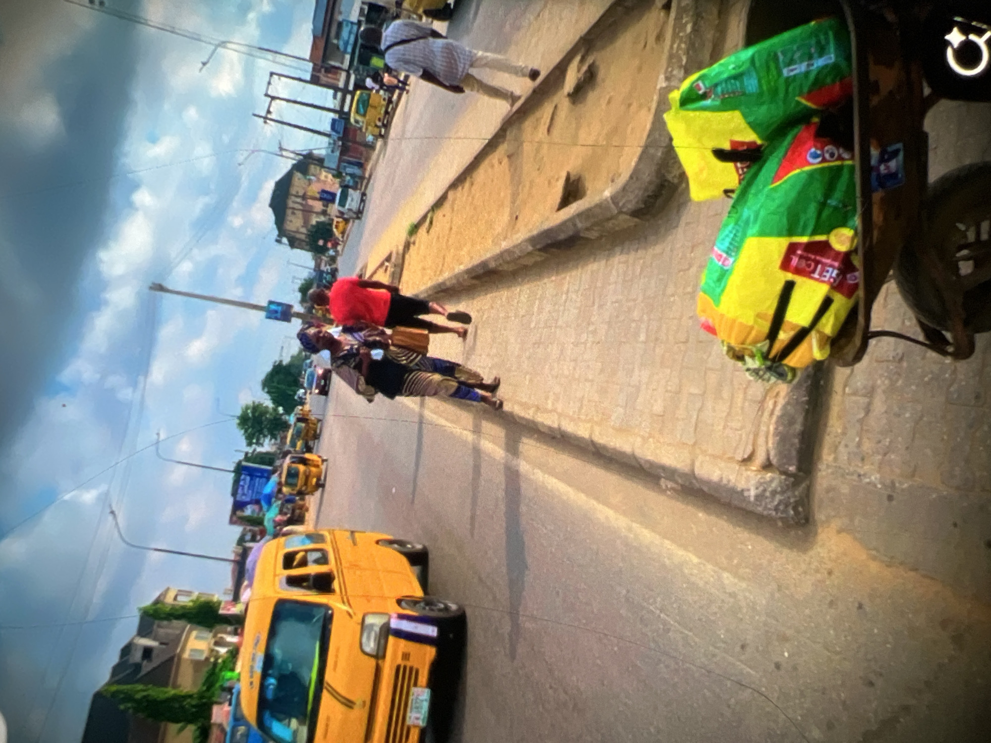 This image captures a street scene in Lagos, Nigeria. A bright yellow commercial vehicle, likely a danfo bus, is parked on the left side of the road. The road is paved, and a raised concrete sidewalk runs alongside it. Several people are visible on the sidewalk and road. Two women, one in a patterned dress and the other in a red shirt and black shorts, are walking along the sidewalk. A man in a light-colored striped shirt and dark pants is walking away from the camera on the right side of the sidewalk. In the foreground, a wheelbarrow filled with bright yellow and green bags is positioned on the ground. The background shows typical urban elements of Lagos, including buildings, streetlights, power lines, and other vehicles. The sky is partly cloudy, with a mix of blue sky and white clouds.
