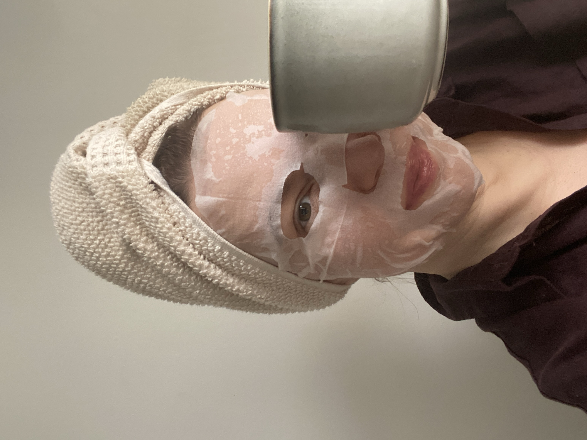 An individual, likely a woman, is depicted engaging in a self-care or relaxation routine indoors. Their face is covered by a white sheet mask, with cutouts revealing the left eye and lips. The visible left eye has a light brown or hazel iris and appears calm. A light-colored, possibly beige or off-white, terrycloth towel is wrapped securely around their head, likely holding back their hair.
A light gray or muted green ceramic mug or cup is positioned close to the person's face, held above the nose and mouth area, suggesting they might be drinking a beverage or using it for a steaming ritual. The person is wearing dark-colored clothing, possibly a maroon or plum-toned shirt or robe, visible around the neck and shoulder area.
The background is a plain, light-colored wall, indicating an indoor setting. The image is taken at a slight angle, with the person's head tilted. The overall atmosphere conveyed is one of tranquility and personal pampering. No specific time of day or weather conditions are discernible from the image. There are no visible textual elements or direct location cues within the frame to confirm the specified location of Chaikovsky, Russia.