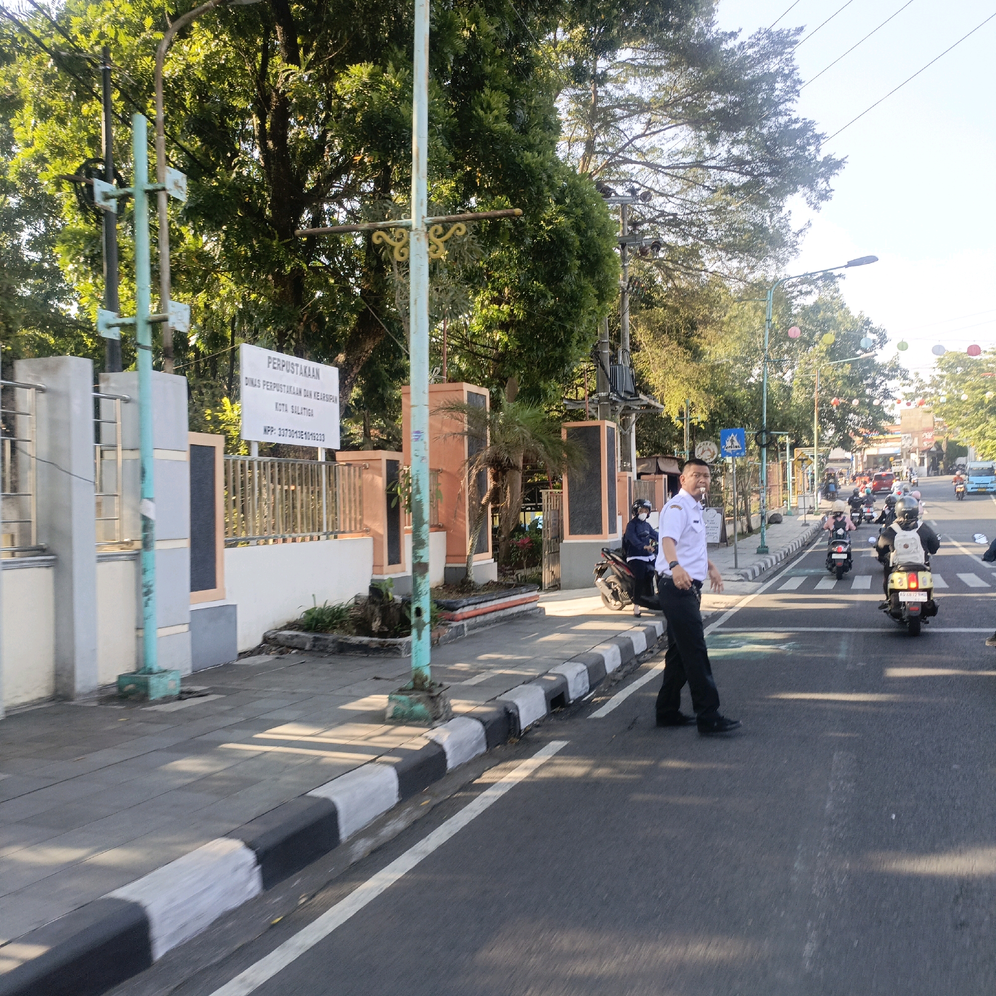 The image shows a street scene in Salatiga, Indonesia, with a pedestrian crossing and several motorbikes. In the foreground, a man in a white shirt and black pants is walking across the street, facing left. He appears to be a traffic officer or uniformed personnel. He is partially obscured by a motorbike with a rider wearing a helmet and a backpack, also crossing the street. Another person in a helmet and dark jacket is visible on a motorbike to the left of the crossing. Behind them, a line of motorbikes is waiting or moving through the intersection. On the left side of the street, there is a sidewalk with a sign that reads "PERPUSTAKAAN DINAS PERPUSTAKAAN DAN KEARSIPAN KOTA SALATIGA" and a registration number. Tall trees with lush green foliage line the street, and power lines are visible overhead. The sky is bright, suggesting it is daytime. Decorative lanterns in various colors are strung across the street in the background. The scene depicts a busy urban environment with moderate traffic.