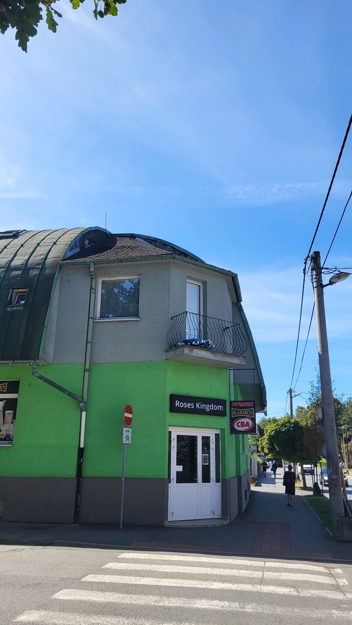 The image shows the corner of a building with a bright green lower story and a gray upper story. The building has a curved, dark-colored roof. A balcony with a decorative railing extends from the upper story. A white door is visible on the front of the building, with a sign above it that reads "Roses Kingdom." To the right of this sign is another sign for "POTRAVINY KARMEN CBA." A street sign with a red circle and a bicycle symbol is posted near the entrance.
A crosswalk is in the foreground, and a sidewalk runs alongside the building. Several trees are visible along the sidewalk. In the background, people are walking on the sidewalk, and a few cars are on the street. The sky is clear and blue. The scene appears to be a street in a town or city, likely during the daytime.
Based on the signs, this location is in Prešov, Slovakia. "Potraviny" means groceries, and "CBA" is a chain of convenience stores in Slovakia. "Karmen" is likely the specific name of the store. "Roses Kingdom" is another establishment present.