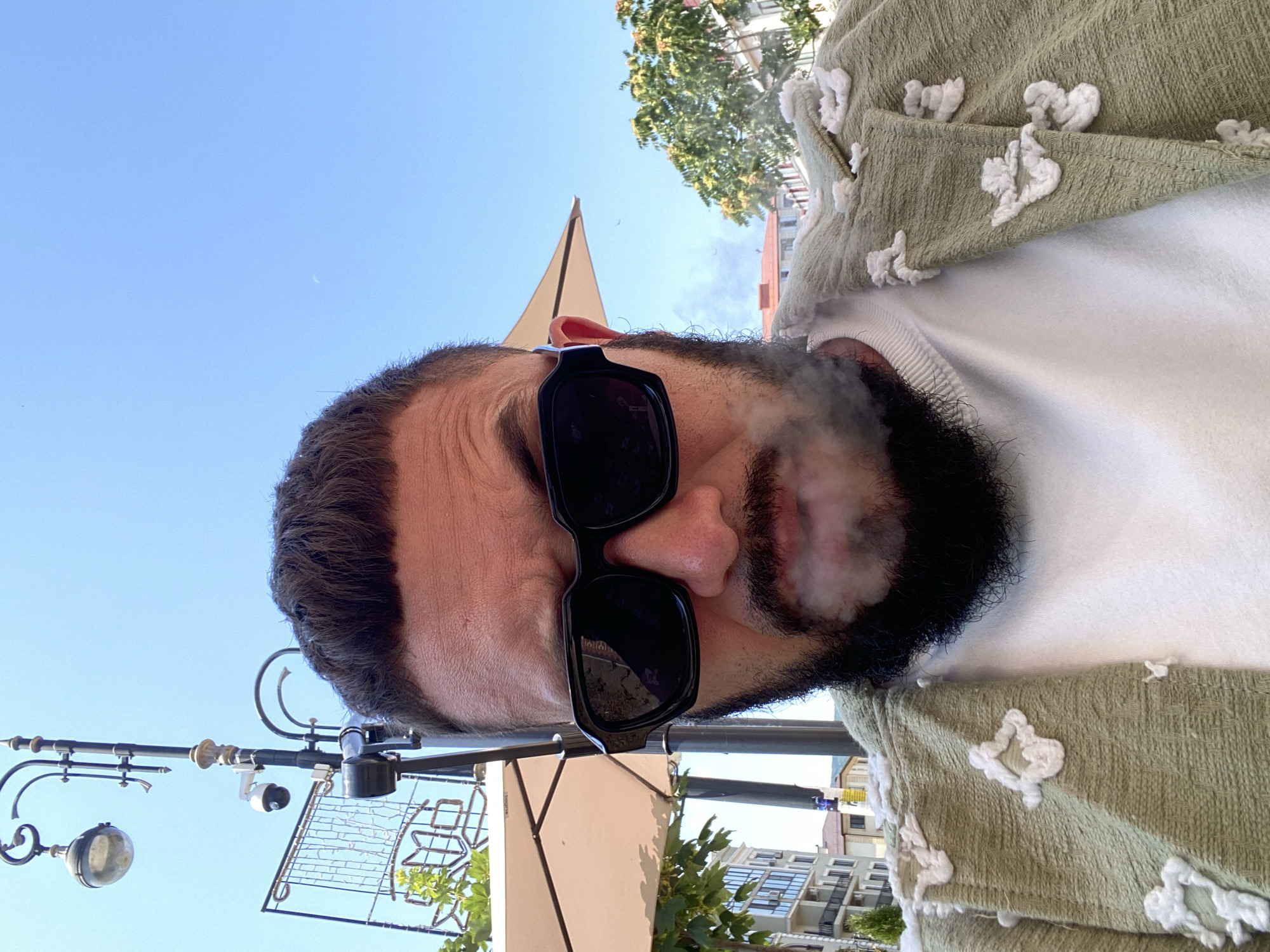 A close-up, selfie-style shot shows a man from the chest up, outdoors during the day. The man is wearing black sunglasses and a light green jacket with white embroidered floral or abstract patterns. A plume of smoke emanates from his mouth, obscuring part of his beard and face. He has dark hair and a full beard. In the background, there is a clear blue sky, a street lamp with a camera attached, a cream-colored umbrella, trees, and buildings characteristic of an urban environment, suggesting the location is a city. The man is positioned centrally in the frame, with the background elements slightly out of focus. The lighting indicates it is daytime.