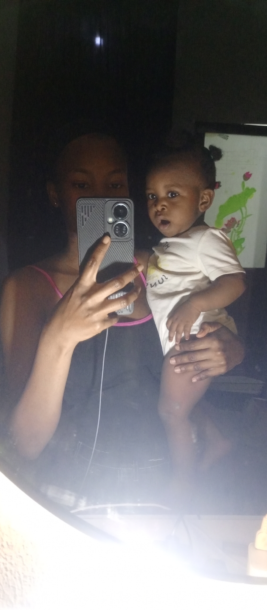 A person holding a baby takes a mirror selfie. The person is wearing a black tank top with pink straps and holding a dark-colored smartphone with a textured case. The baby is wearing a white onesie. The background is dark but includes a picture with green and pink elements. The location appears to be inside a room, city and specific region not determined.