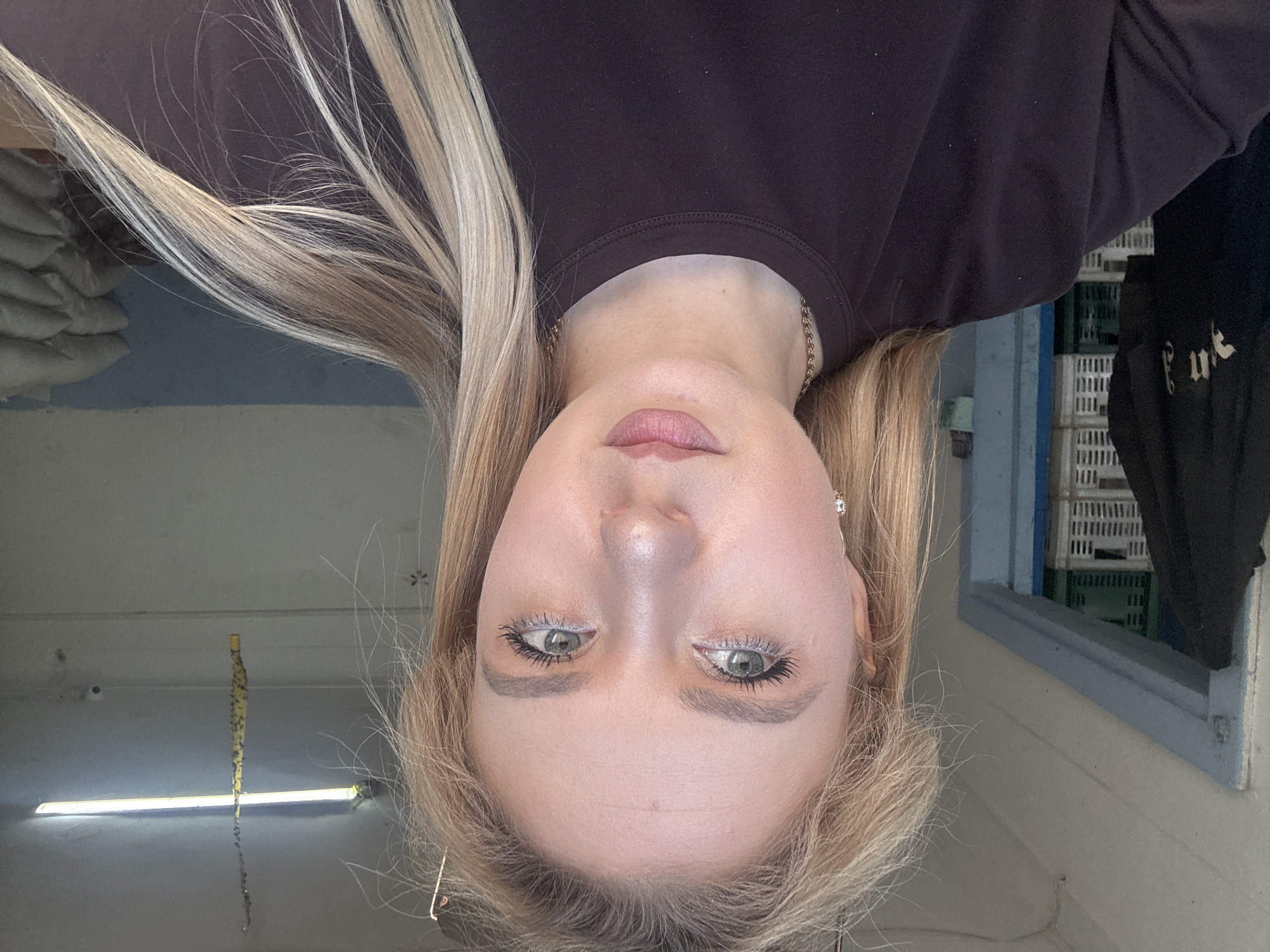 A young woman with blonde hair, wearing a dark brown t-shirt, is captured in a selfie. She is positioned centrally in the frame, with her face slightly tilted. Her eyes are a light green, and she is wearing subtle makeup, including mascara and lipstick. A delicate silver chain necklace is visible around her neck. The background reveals an interior setting. To her left, a dark jacket hangs on a door frame, with white plastic crates visible behind it. The environment appears to be dimly lit, with fluorescent lighting visible on the ceiling. In the background to the right, stacks of what appear to be large sacks are discernible.