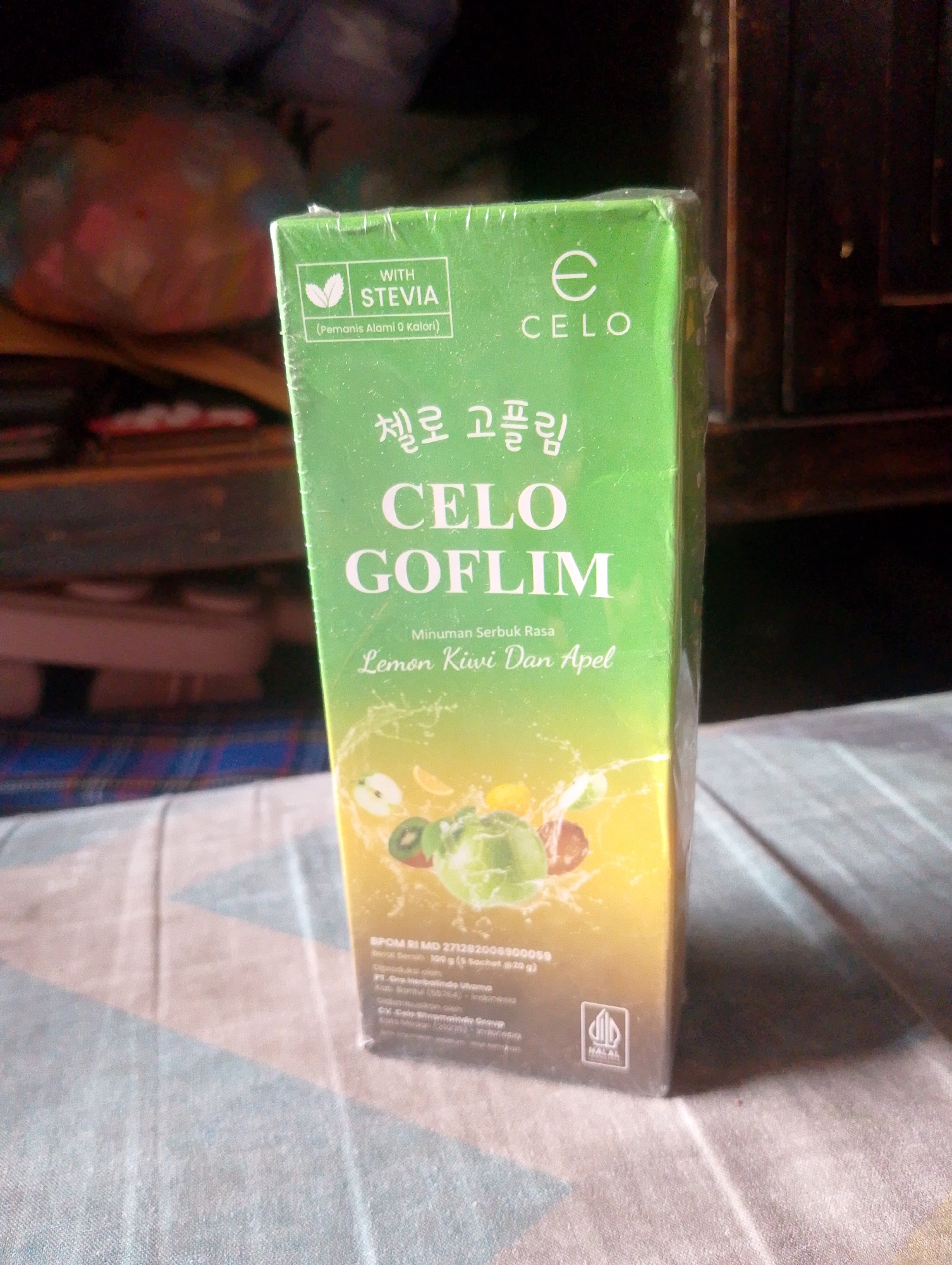 An eye-level, medium shot captures a brightly lit green box of "CELO GOFLIM" from the front. The box is wrapped in cellophane and rests on a patterned cloth. The background is dark and out of focus, suggesting an indoor setting. The box features Korean text and English text indicating "WITH STEVIA" and "Pemanis Alami 0 Kalori" (Natural Sweetener 0 Calories). The bottom of the box has Indonesian text, including "BPOM RI MD 271283000059" and "Net Weight: 168 g (8 Sachets @ 21 g)". A Halal logo is visible on the bottom right corner of the box.