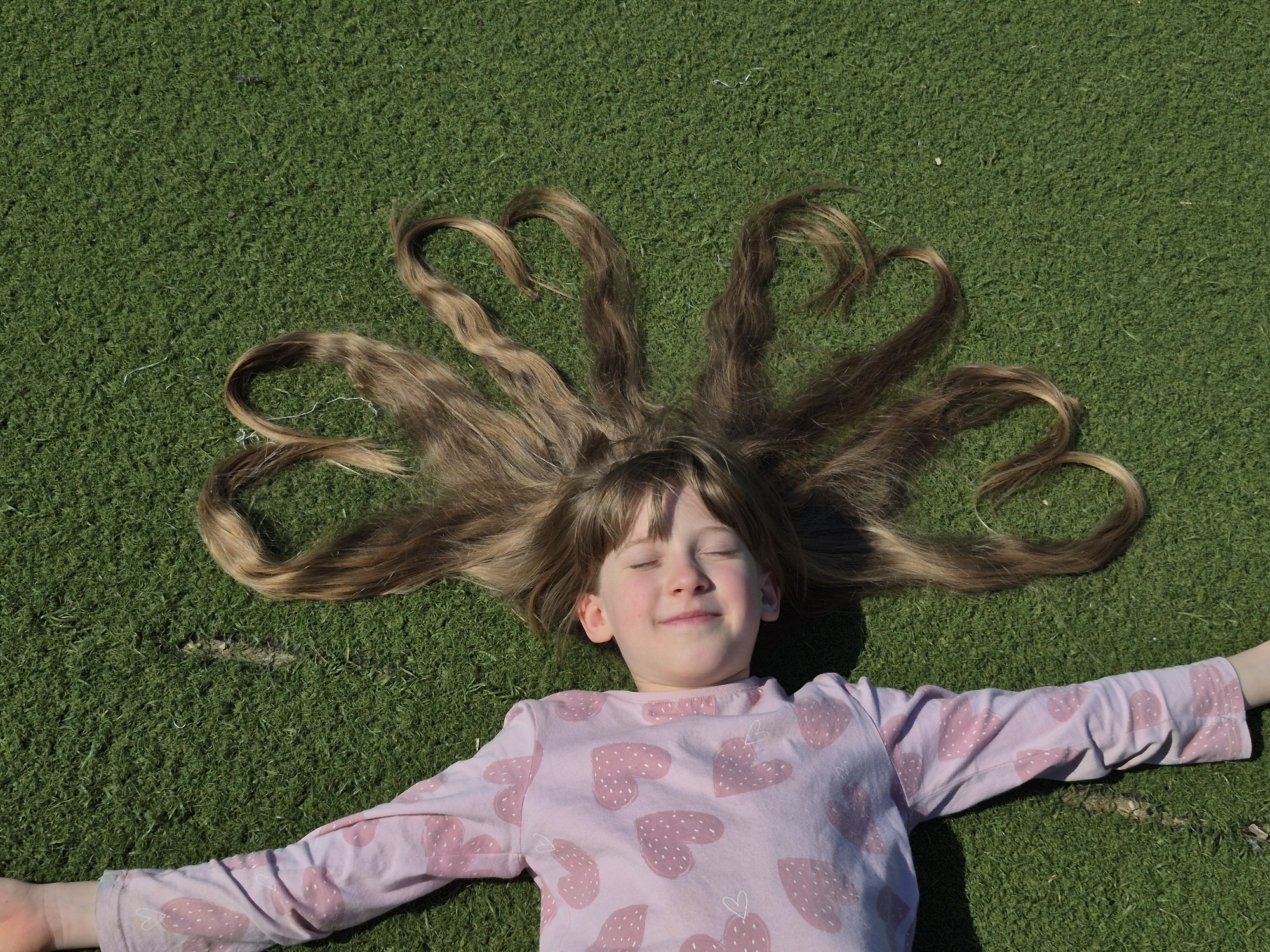 A girl lies on artificial grass. Her arms are spread out. She is wearing a pink, long-sleeved shirt with pink heart shapes printed on it. Her eyes are closed. Her long, brown hair is spread out around her head. The ends of several strands of her hair are shaped into hearts. This photograph was likely taken in Chaikovsky, Russia.