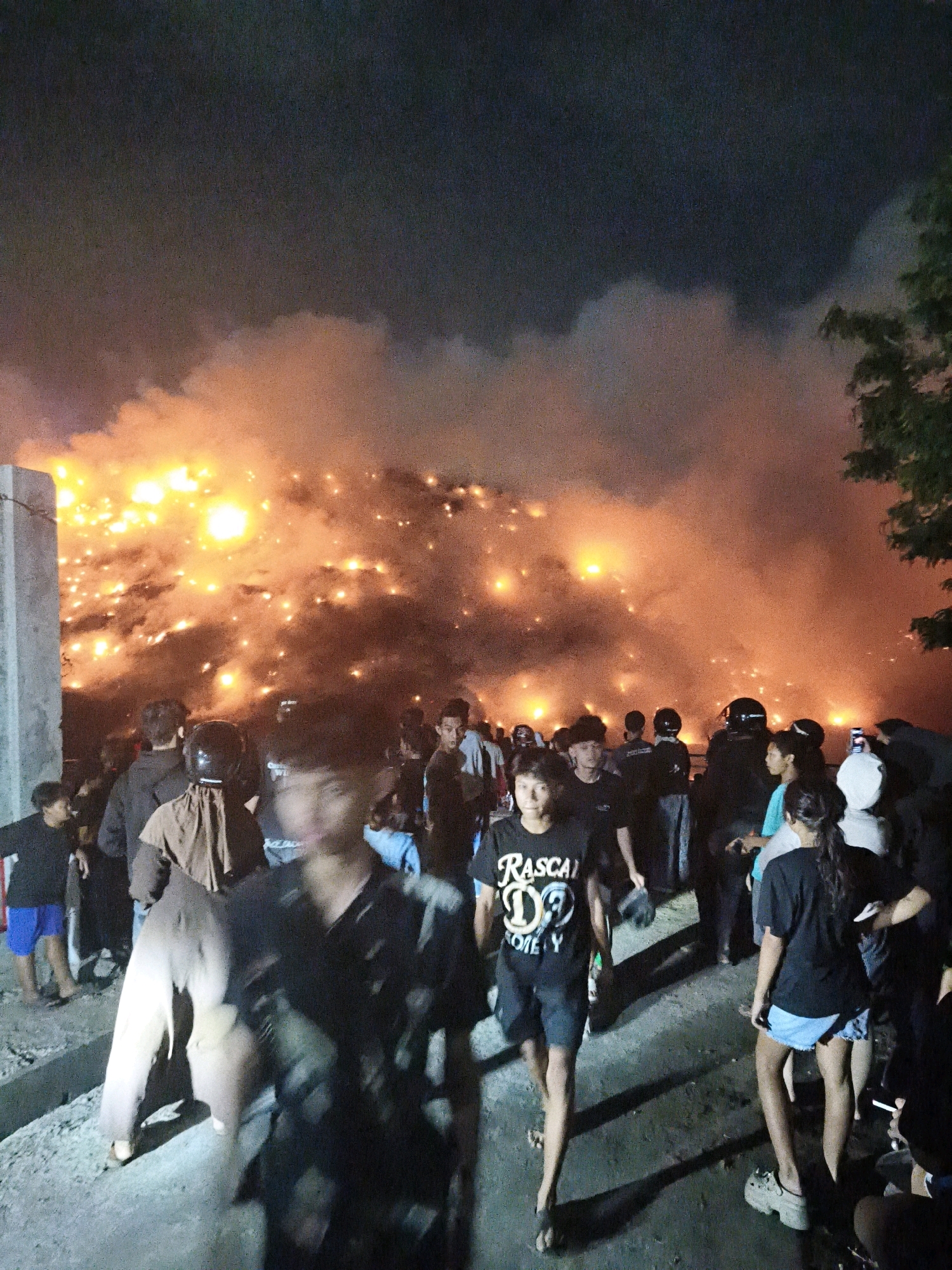 An outdoor scene at night in Surakarta, Indonesia, depicts a crowd of people observing a large, intense fire that is illuminating the sky with bright orange and yellow flames and smoke. The fire appears to be emanating from a pile of material, possibly waste or debris, situated behind the onlookers. The crowd consists of individuals of various ages, some are wearing casual clothing, while others have on protective gear such as helmets, suggesting a possible organized event or response. The ground is uneven and appears to be a dirt or gravel path. A concrete pillar is visible on the left side of the image. The overall atmosphere is chaotic and dramatic due to the large fire and the gathering of people.