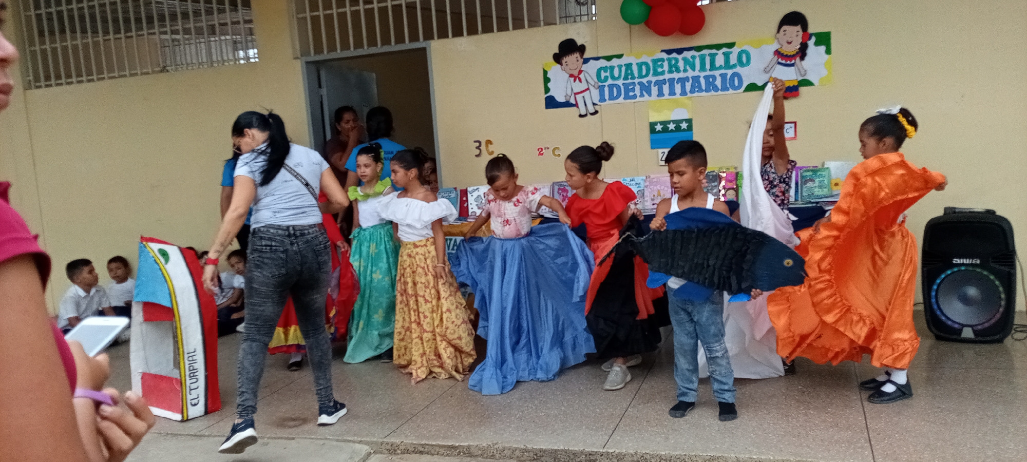The image shows a group of children and adults in what appears to be a school setting in the city of Unknown, Unknown. The children are dressed in colorful traditional costumes. A boy holds a large paper mache fish. Some adults are present, seemingly supervising the children. The setting is in front of a building with a poster on the wall that reads "Cuadernillo Identitario". A black speaker is seen on the right.