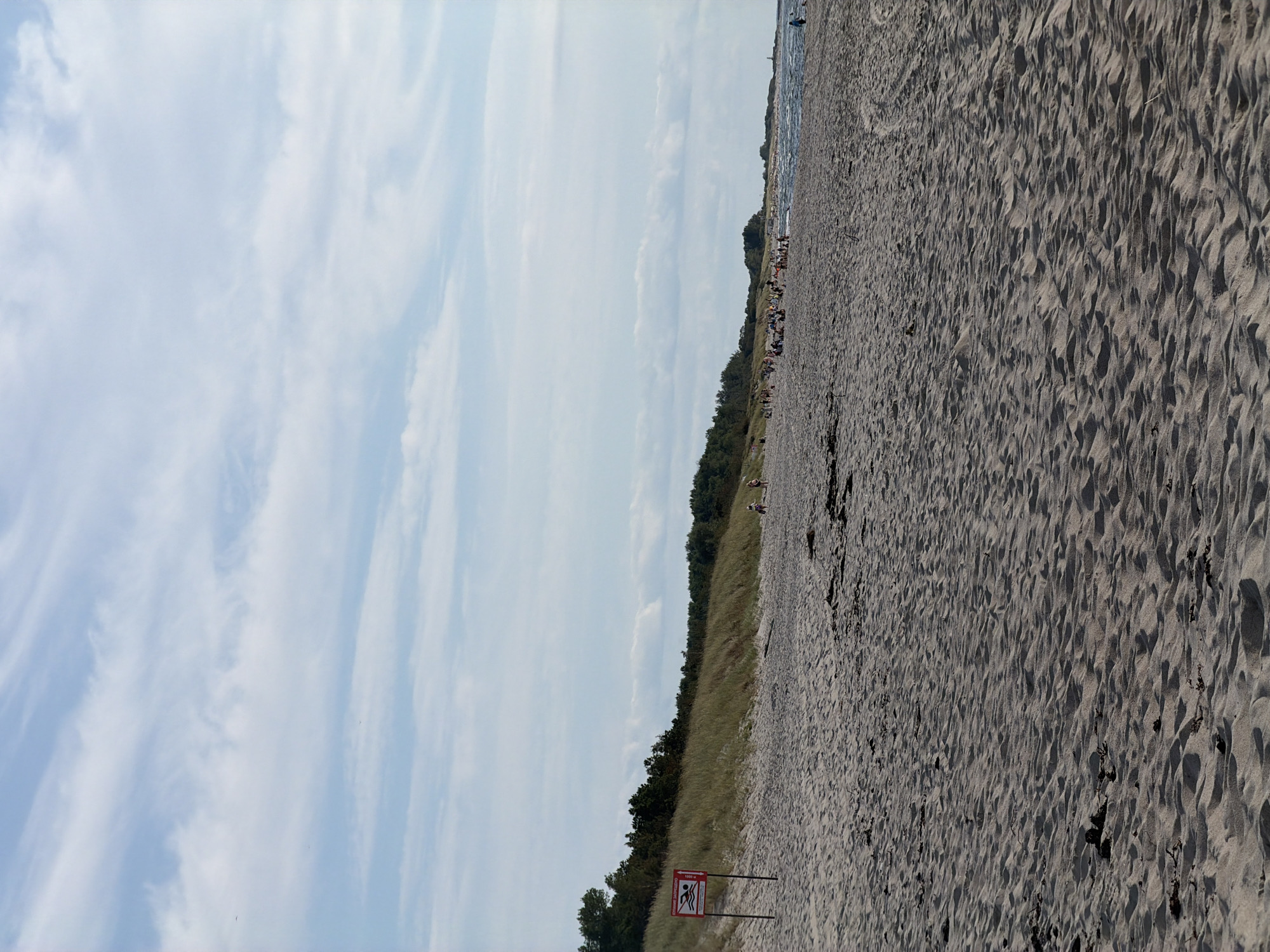 The image depicts a wide, sandy beach in Baltiysk, Russia, under a partly cloudy sky. The foreground is dominated by the texture of the sand, showing ripples and shadows from the sunlight. In the middle ground, a line of vegetation, consisting of grass and trees, forms a backdrop to the beach. A red and white sign with a symbol indicating swimming is prohibited is visible on the left side of the beach. Scattered along the shoreline and further down the beach, several individuals and small groups of people are present, some appear to be sunbathing or walking. The sea is visible in the distance, with gentle waves and a lighthouse or tower in the far right background.