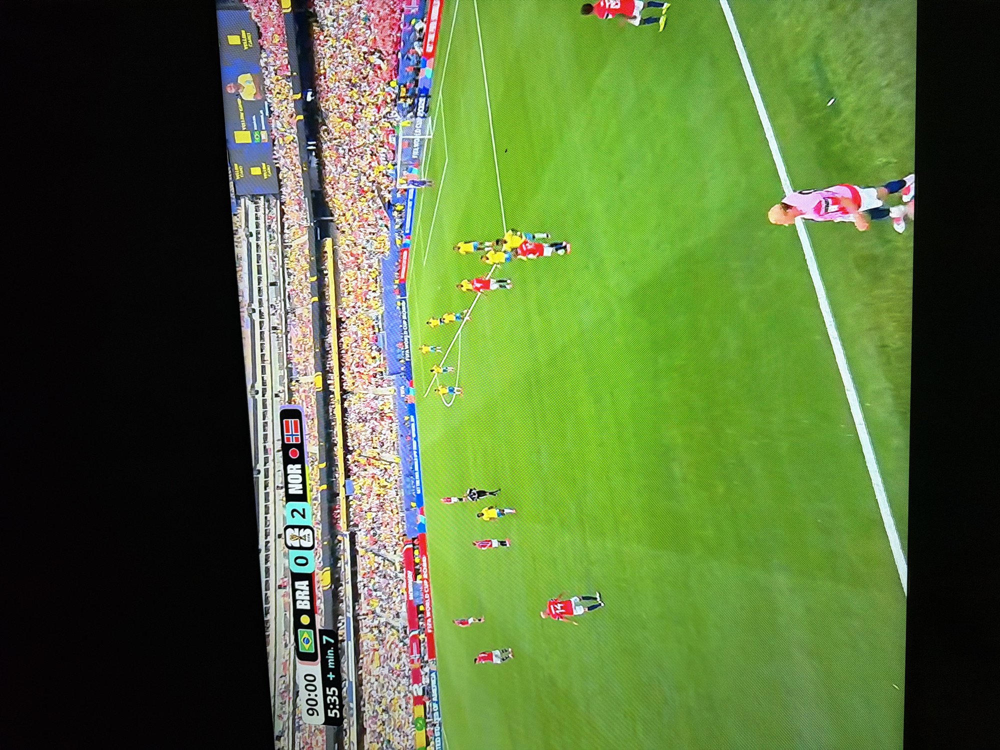 The image captures a live broadcast of a soccer match. A scoreboard at the top displays "90:00" indicating the current game time, followed by the score "BRA 0 2 NOR". Below that, it reads "5:35 + min. 7". The soccer field is visible with players in red and yellow/green uniforms. A player in a red uniform with the number 14 is in the foreground. In the background, spectators are seated in the stadium stands. The image does not provide any information about the location in Ognianovo, Bulgaria.