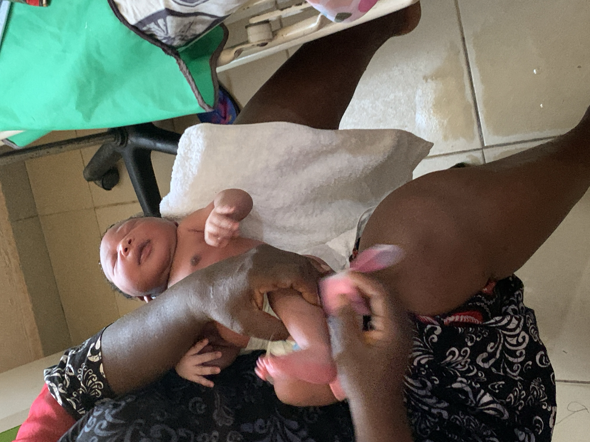 A newborn infant is being held in the arms of an adult, who is sitting on the floor. The infant is lying on a white towel and has a light-colored complexion. The adult has dark skin and is wearing a black and white patterned garment. The location appears to be indoors, on a tiled floor. The location may be in Jos, Nigeria, but that is difficult to confirm from the image.