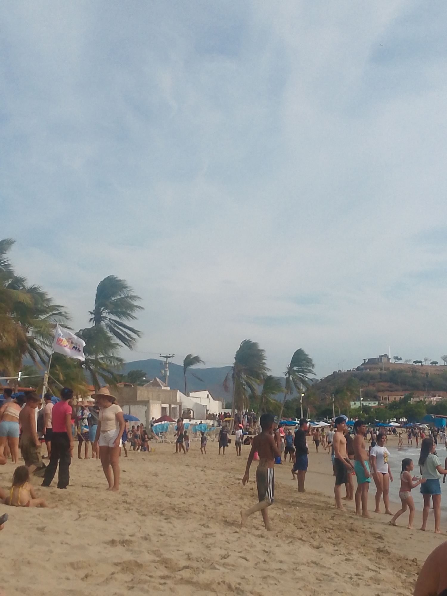 The image shows a crowded beach scene, likely in Juan Griego, Venezuela, based on the presence of a hill with buildings and a white monument, which is characteristic of the location. A multitude of people are on the sandy beach, including children and adults. Some are wearing bathing suits and casual clothing. Some people are walking towards the water, while others are sitting or standing. Palm trees are visible along the beach, some of them swaying in the wind. A flag is seen near the center of the image. The sky is partially cloudy. In the background, a hill is visible with buildings and vegetation on it.