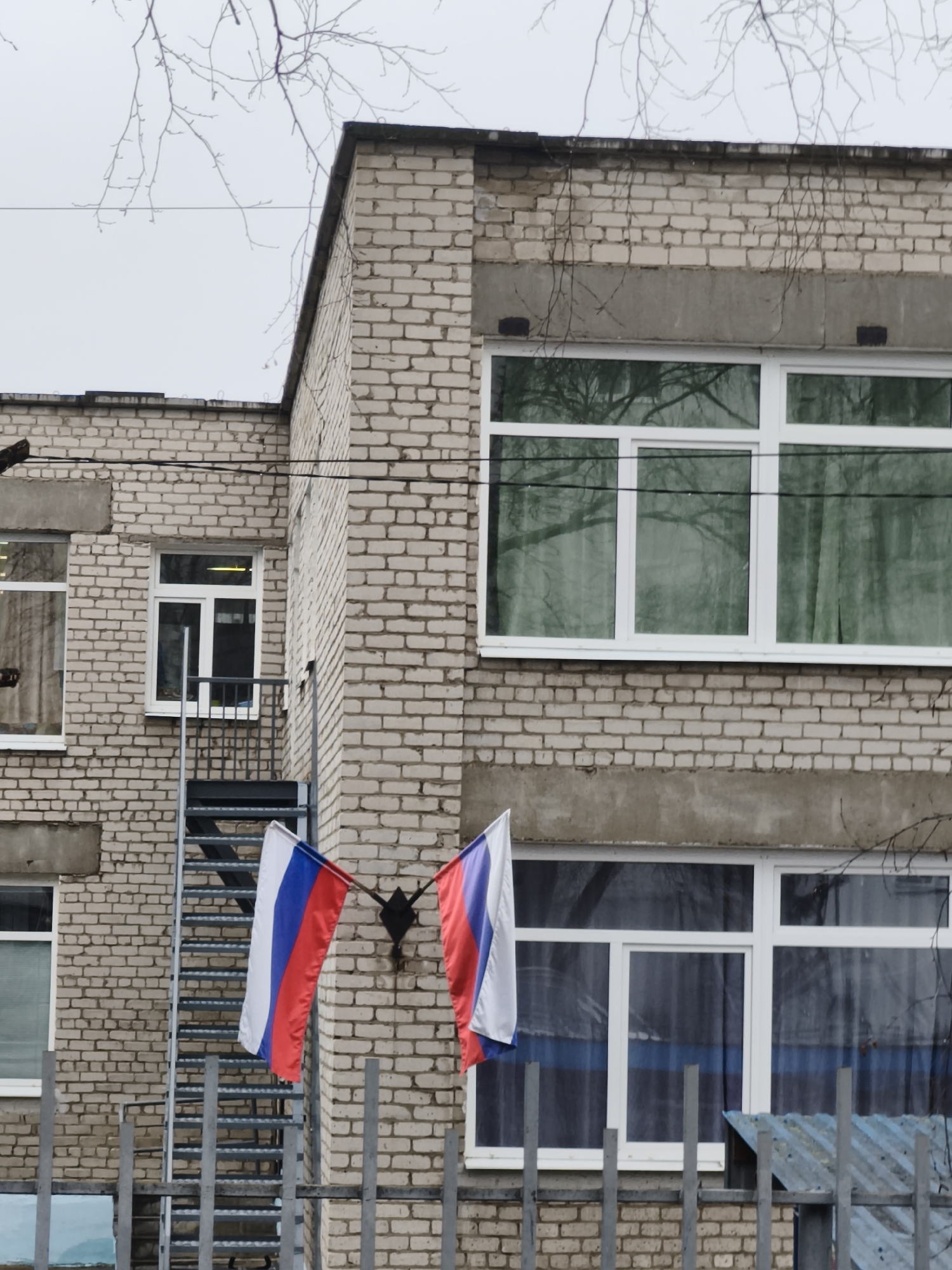 The image captures an exterior view of a light-colored brick building, likely a multi-story structure, under an overcast sky. The building features several windows with white frames; some reflect bare tree branches, while others show glimpses of interior curtains, including one with a blue horizontal stripe.
Prominently displayed on the building's facade are two small Russian flags, with horizontal white, blue, and red stripes, hanging from a fixture. An external metal staircase, possibly a fire escape, is visible on the left side of the building, leading up to a smaller window.
In the foreground, a metal fence with vertical bars partially obscures the lower section of the building. The sky is a uniform, pale gray, indicating an overcast day. Bare tree branches, devoid of leaves, are visible against the sky and reflected in some windows, suggesting a late autumn or winter season. Several black electrical wires traverse horizontally across the mid-ground of the scene. No people or visible text are present in the image.