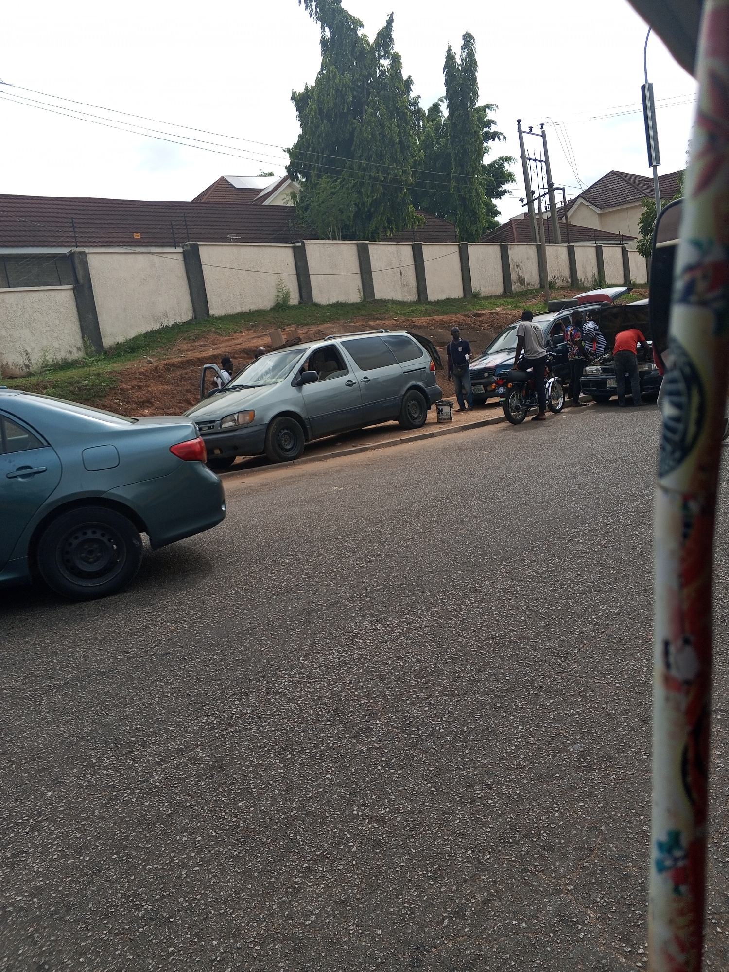 The image shows a street scene in Abuja, Nigeria. Several vehicles are parked along the side of the road. A blue sedan set back, a grey minivan, a dark-colored sedan, and potentially other partially obscured vehicles are present. Several people are gathered near the dark-colored sedan, possibly working on the car, with the hood open. One person is standing near a motorcycle parked beside the sedan. There is a wall separating the street from a property with a house and trees in the background. A pole with designs is in the foreground.