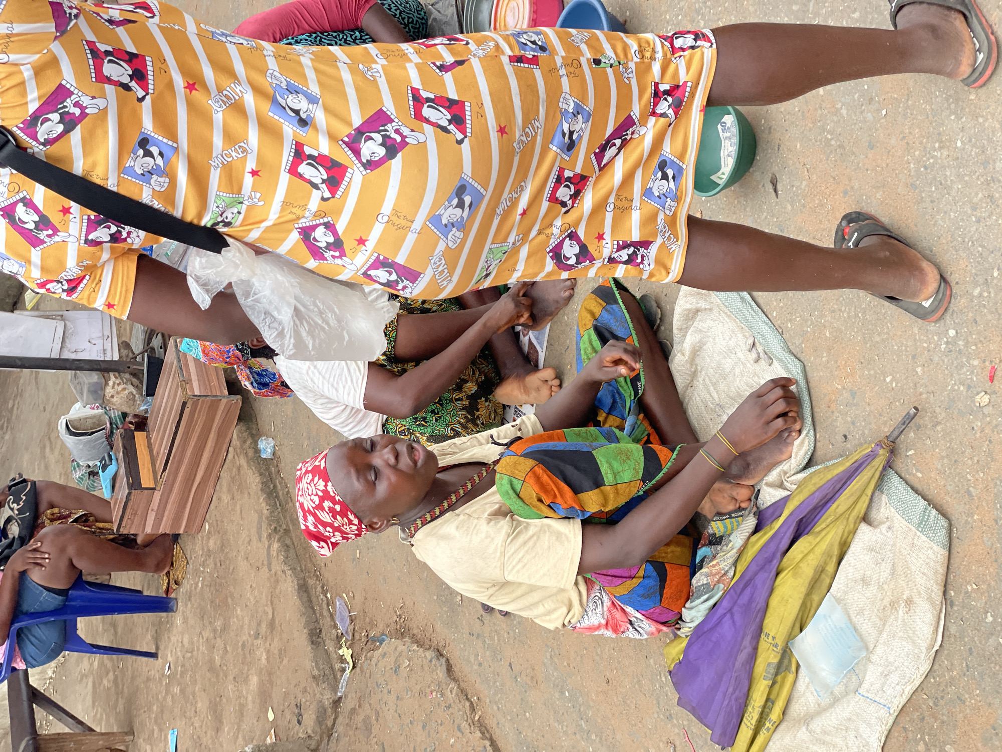 The image shows an outdoor scene, likely a market or street in Onitsha, Nigeria. The ground is a mix of concrete and dirt. The dominant figures are several people, most likely women. One woman is sitting on the ground with her legs crossed, wearing a light-colored top and a colorful, patterned skirt. She also has a red headscarf. Another woman is standing, wearing a yellow dress with a Mickey Mouse pattern. A third woman is sitting on a blue plastic chair. There are also bags and objects scattered around, including a folded umbrella. A partially visible wooden structure is also visible.