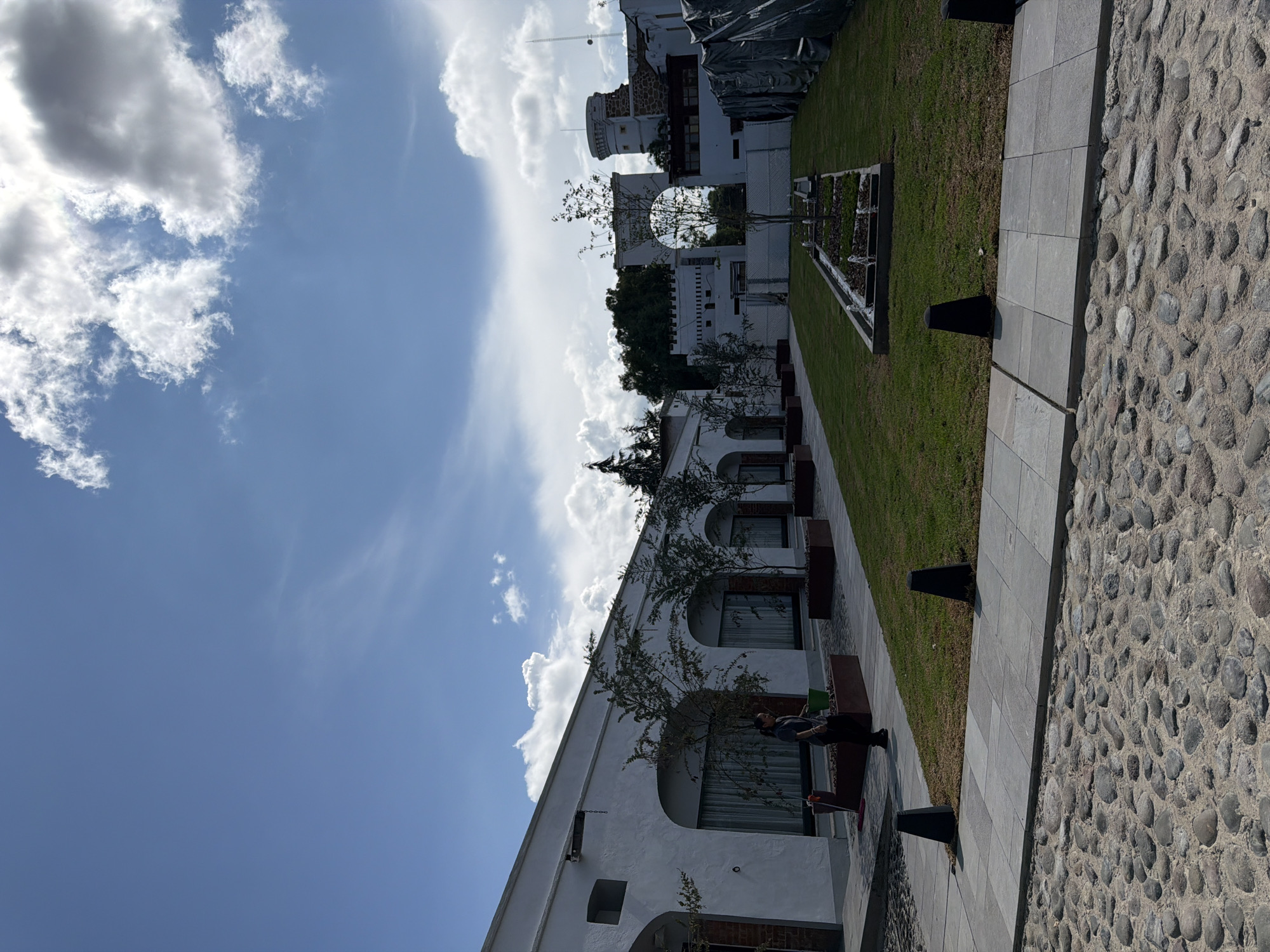 This image captures an outdoor scene in what appears to be a public or private courtyard, possibly in San Lucas el Grande, Mexico, given the architectural style. The scene is dominated by a clear blue sky with numerous fluffy white clouds.
On the left side of the frame, several white buildings with arched windows and dark frames are visible. Small, young trees are planted in rectangular planters along the base of these buildings, offering a touch of greenery against the white walls. One person is visible, sitting on a dark bench positioned in front of one of the buildings, appearing to relax. Further back, another white building features a prominent circular opening or archway, with a darker, possibly older structure behind it, partially covered by what looks like scaffolding or protective tarps. A small tower-like structure is also visible in the distance.
The right side of the image features a well-maintained green lawn, bordered by a light gray paved walkway made of square tiles. Adjacent to this walkway is a textured cobblestone path or area, composed of rounded, smooth stones. A long, rectangular planter box with small plants is embedded in the lawn further back. Several dark, triangular or pyramidal bollards are spaced along the edges of the paved areas.
The time of day appears to be daytime, with bright sunlight casting shadows, suggesting mid-morning or afternoon. The overall atmosphere is peaceful and serene. There is no visible text in the image.