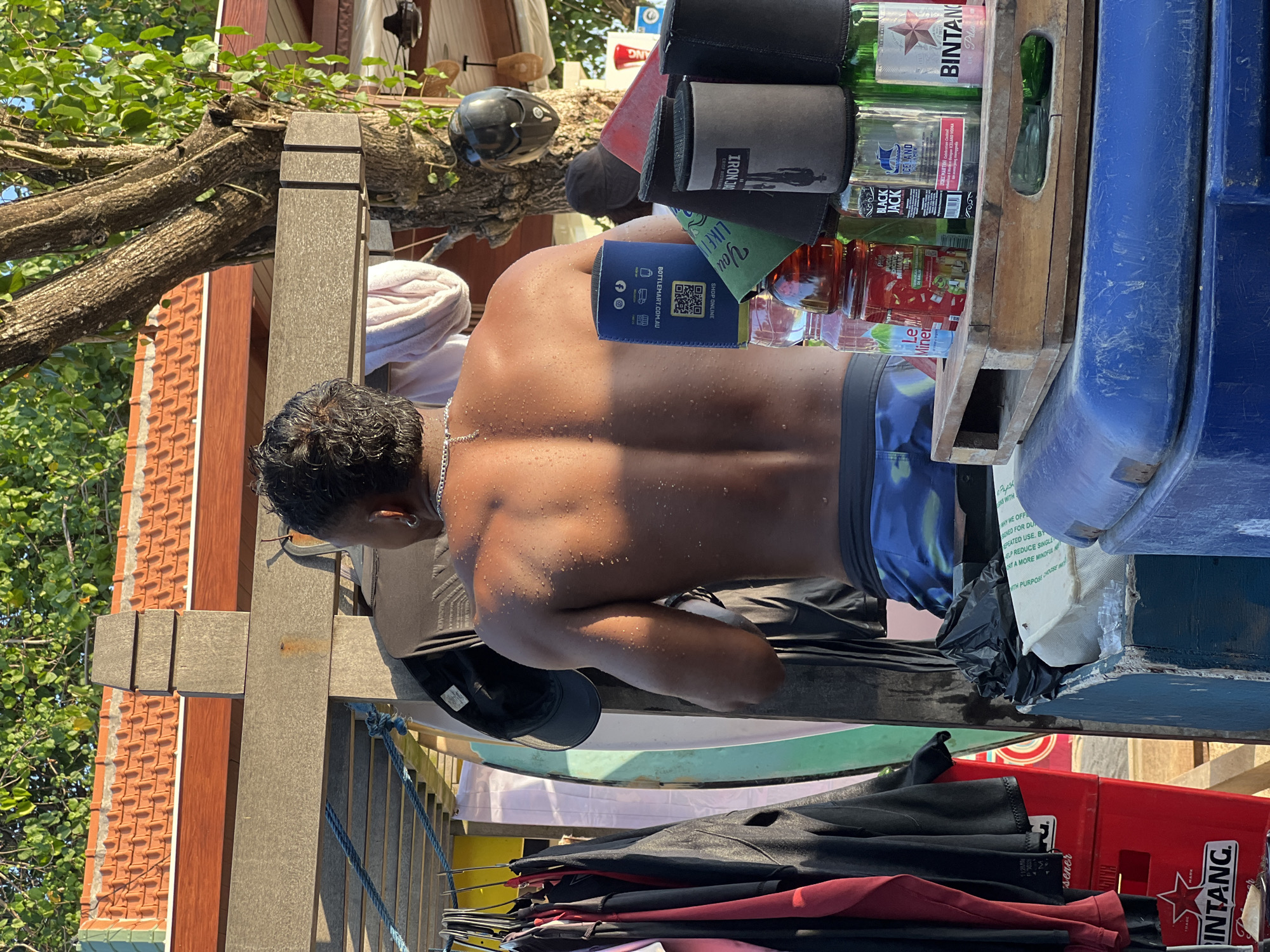 In Kuta, Indonesia, on a sunny day, a person with wet skin and dark, curly hair stands with their back to the camera. They are wearing blue patterned swim trunks and a silver chain necklace. Their shoulders and back are visible, glistening with water droplets. Behind them, a rack holds various clothing items, including t-shirts and what appear to be wetsuits, along with surfboards. A wooden structure supports the rack and clothing. In the foreground, several bottles, including Bintang beer and what appears to be a flavored beverage, are placed on a wooden tray atop a blue cooler. A red crate with the Bintang logo is also visible to the left. The background shows trees, a tiled roof, and parts of a building, suggesting an outdoor setting likely near a beach or tourist area.