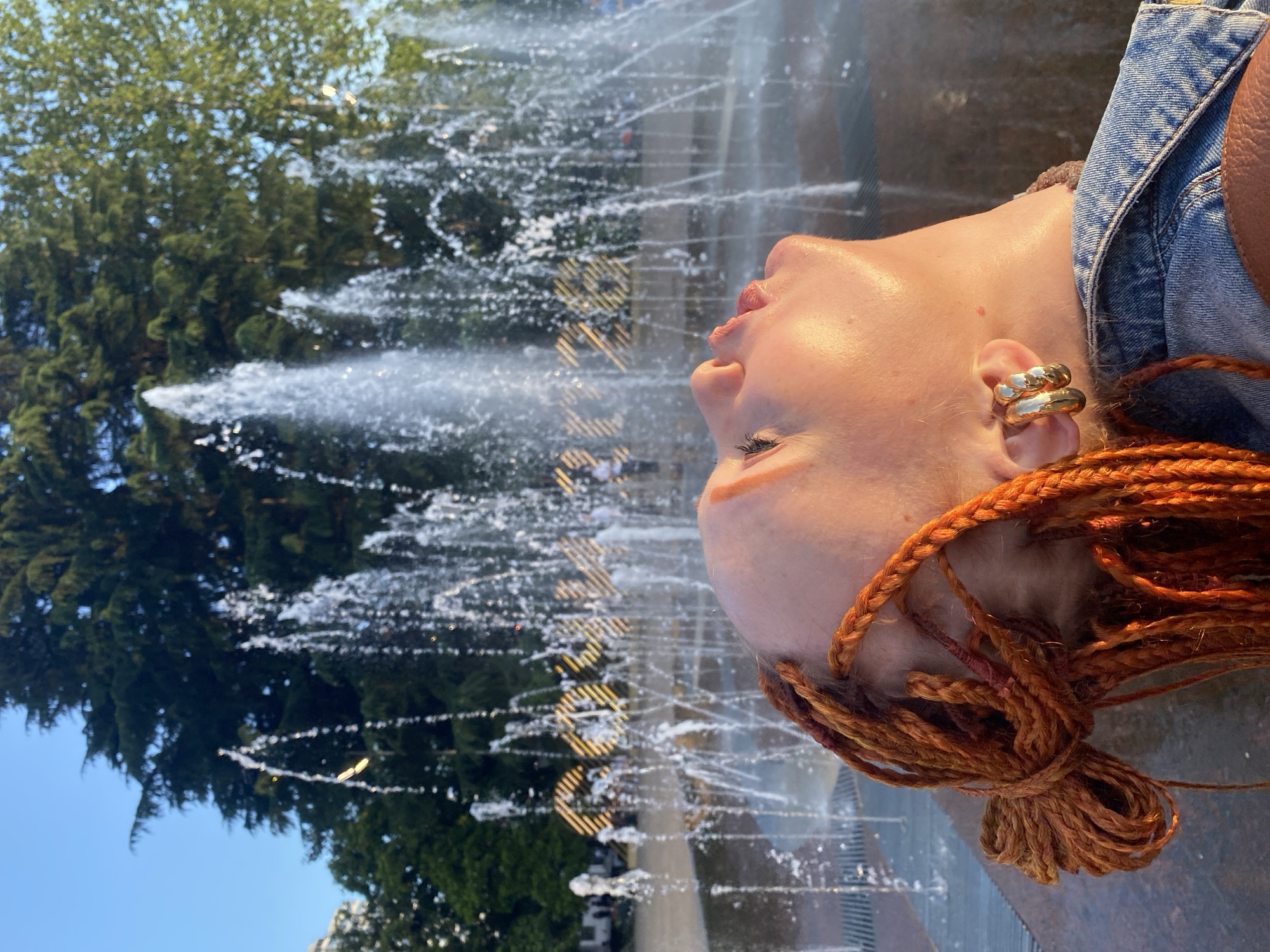 A close-up, low-angle shot shows the side profile of a young person with copper-colored braids, tilting their head back towards a fountain in the city of Sochi, Russia. The setting sun casts a warm glow on the person's face, highlighting their features, including closed eyes, a slightly open mouth, and a prominent gold hoop earring. The person is wearing a denim jacket. The fountain features multiple jets of water spraying upwards, creating a misty effect, and in the background, large, illuminated golden letters spell out "SOCHI 2024". Trees and a clear blue sky complete the scene.