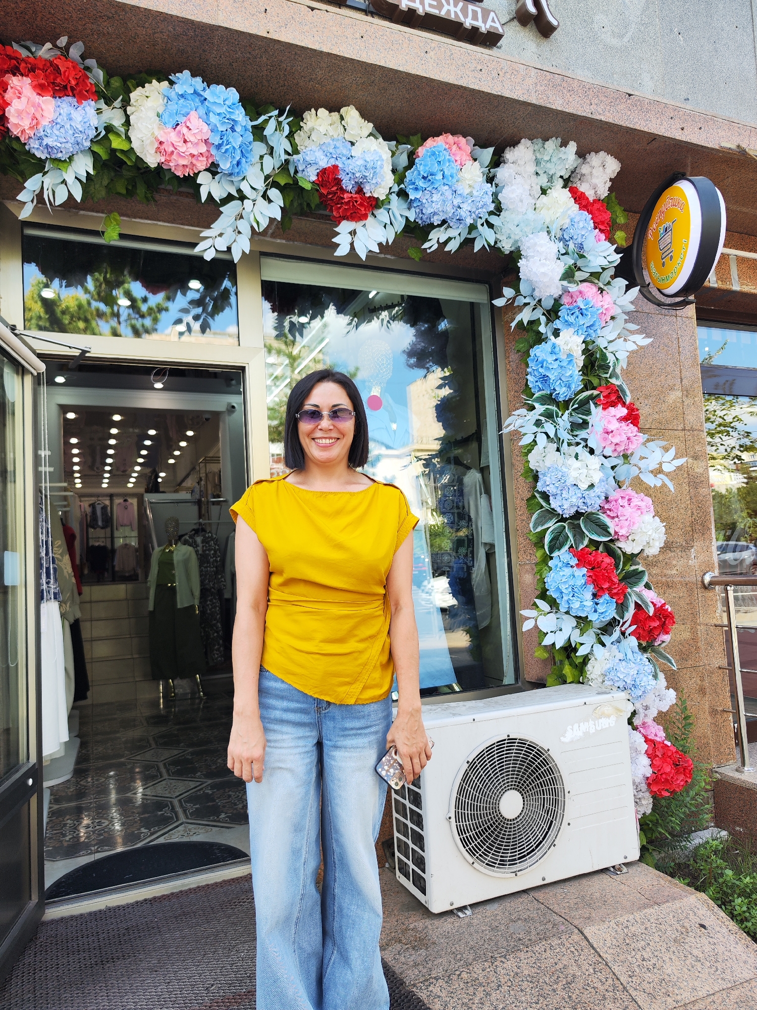 A woman with dark hair and sunglasses stands smiling in front of a store adorned with artificial floral decorations. She is wearing a yellow, asymmetrical top and light blue flared jeans. To her right, an outdoor air conditioning unit sits on the ground. Behind her, the entrance of the store, labeled "Одежда" (Clothing), is visible, displaying mannequins and racks of clothes. The store's facade is made of granite. The scene appears to be on a street, with trees and reflections of the sky visible in the store's windows. The store sign also features a circular, illuminated advertisement with Cyrillic text and a shopping cart icon, indicative of a retail establishment. The presence of "Одежда" on the sign and the overall setting suggest the location is a clothing store in Astana, Kazakhstan.