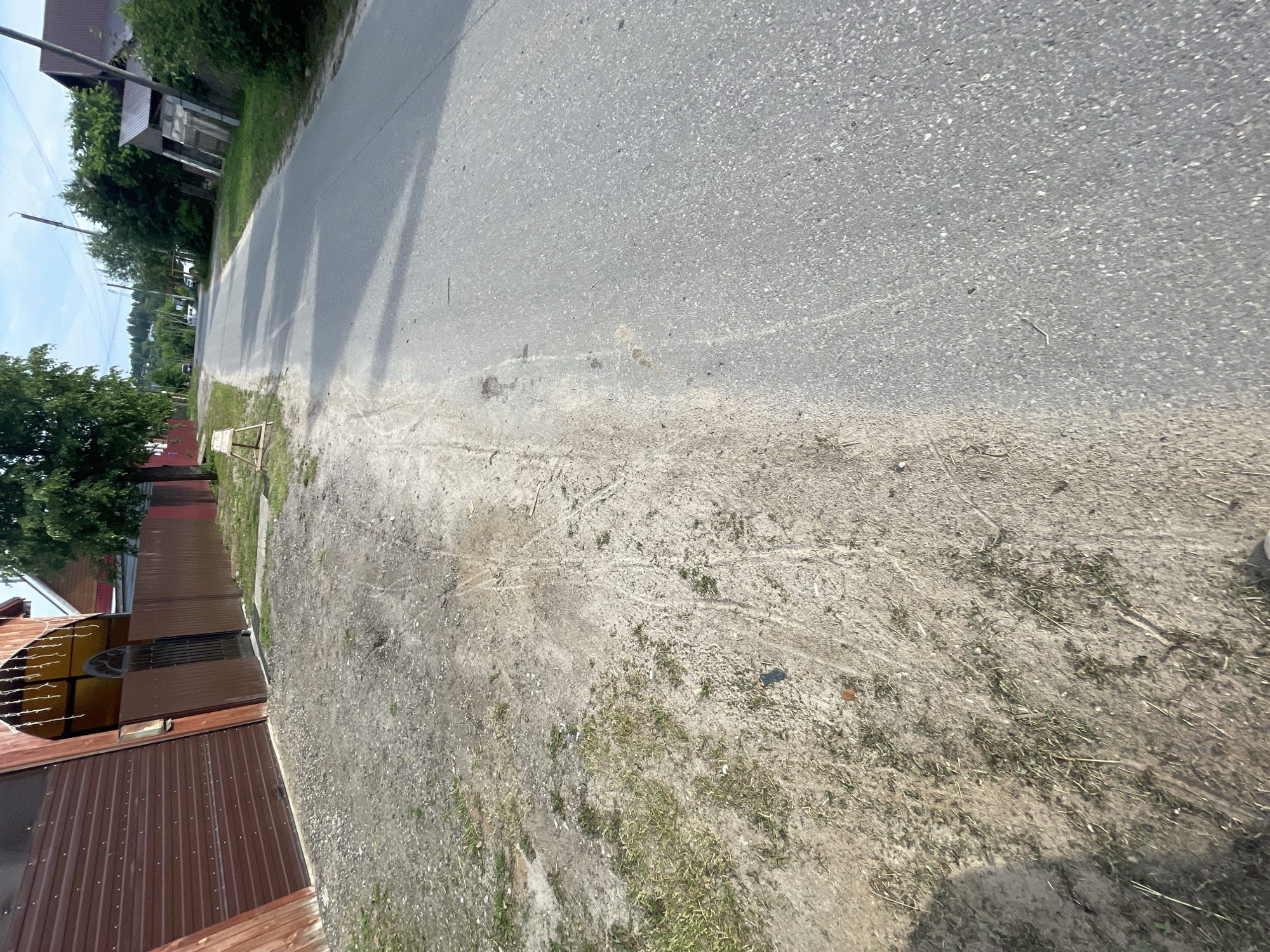 The image shows a street in Krasnye Baki, Russia, on a sunny day. The street is paved with asphalt and has a dirt shoulder on the left side. A brown metal fence runs along the side of the road, with a large brown gate leading to a building with a red roof. A wooden bench is also visible on the dirt shoulder. Trees and houses are seen in the background. The scene appears to be a residential area.