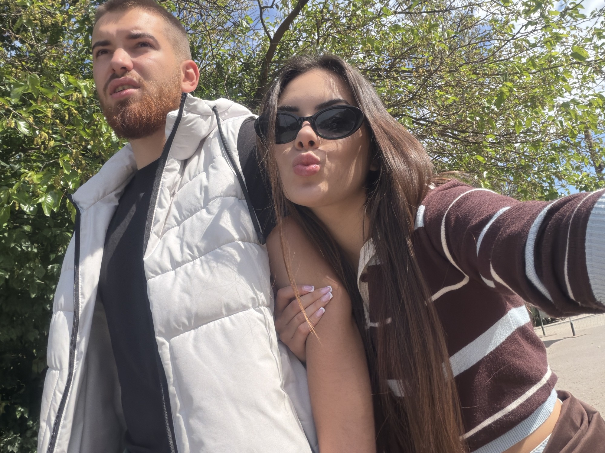 The image shows a man and a woman in an outdoor setting, likely in Sofia, Bulgaria, based on the presence of trees and urban features. The man, on the left, has short brown hair, a beard, and is wearing a black t-shirt under a white puffy jacket. He is looking off to the side. The woman, on the right, has long brown hair and is wearing sunglasses and a brown and white striped sweater. She is making a kissing face towards the camera and has her arm around the man. The background includes trees and a glimpse of a paved surface, suggesting they might be in a park or along a street.