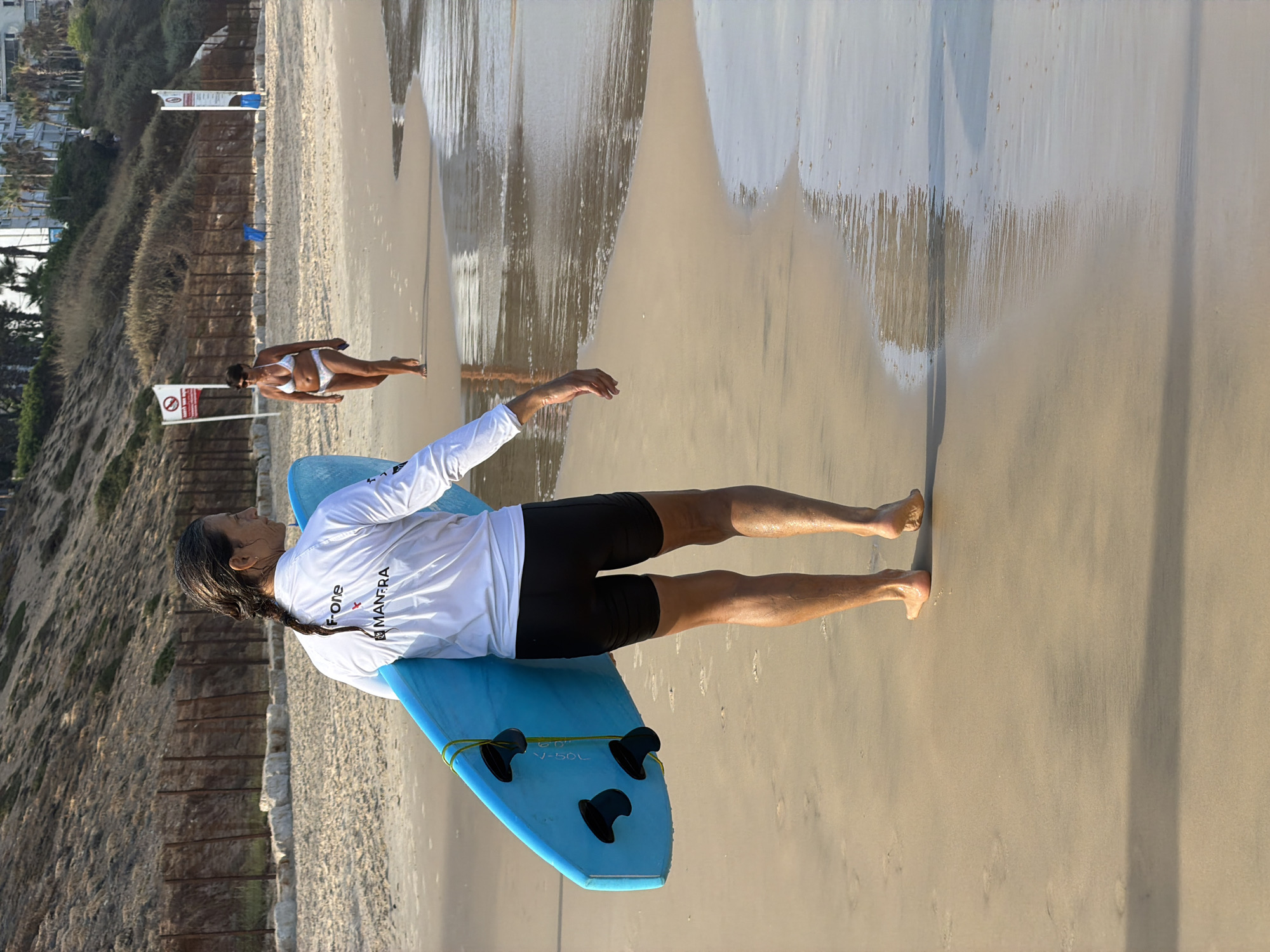 The image shows a beach in Bat Yam, Israel. In the foreground, a person with dark hair in a braid walks away from the camera, carrying a blue surfboard. They are wearing a white long-sleeved shirt with logos on the back and black shorts. The person's legs and feet are wet from the shallow water. Further back on the beach, a woman in a white bikini walks towards the water. The beach has sand and some rocks, with a fence line and vegetation on a slope behind it. The ocean is visible with gentle waves washing onto the shore. There are some signs visible in the background.