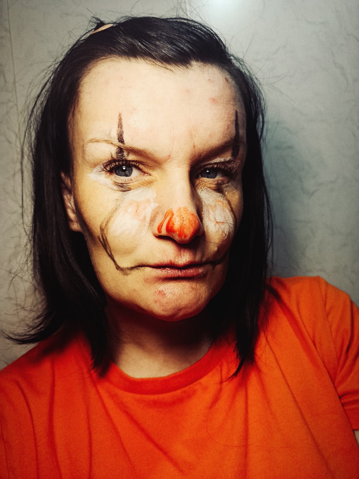 A medium shot shows a person wearing an orange shirt, their face painted in clown makeup. The makeup includes a red nose, white patches around the eyes and nose, and black lines extending from the corners of the eyes and mouth. There's also a vertical black line painted on the forehead. The person's gaze is directed toward the camera with a somewhat cynical expression. The background appears to be a plain, light-colored wall. There is no indication in the image that the location is Фоки, Russia.