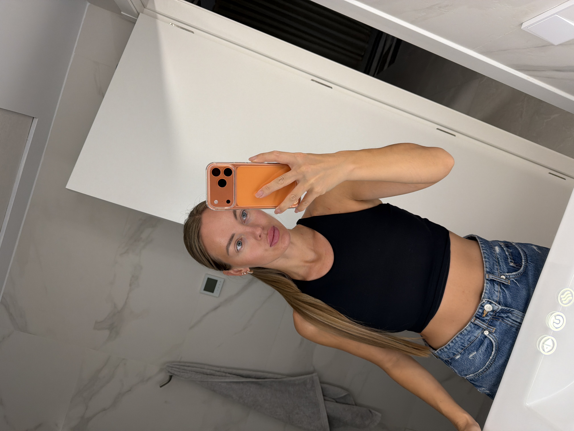 A blonde woman is taking a selfie in a bathroom, holding an orange-cased smartphone in her right hand. She is wearing a black crop top and blue denim jeans. Her hair is pulled back into a ponytail, and she is wearing a gold hoop earring in her visible left ear. The bathroom features a marble-patterned wall, a grey towel hanging on a hook, and a white sink. The overall lighting is dim. The scene does not provide any specific indicators of its location within Finestrat, Spain.