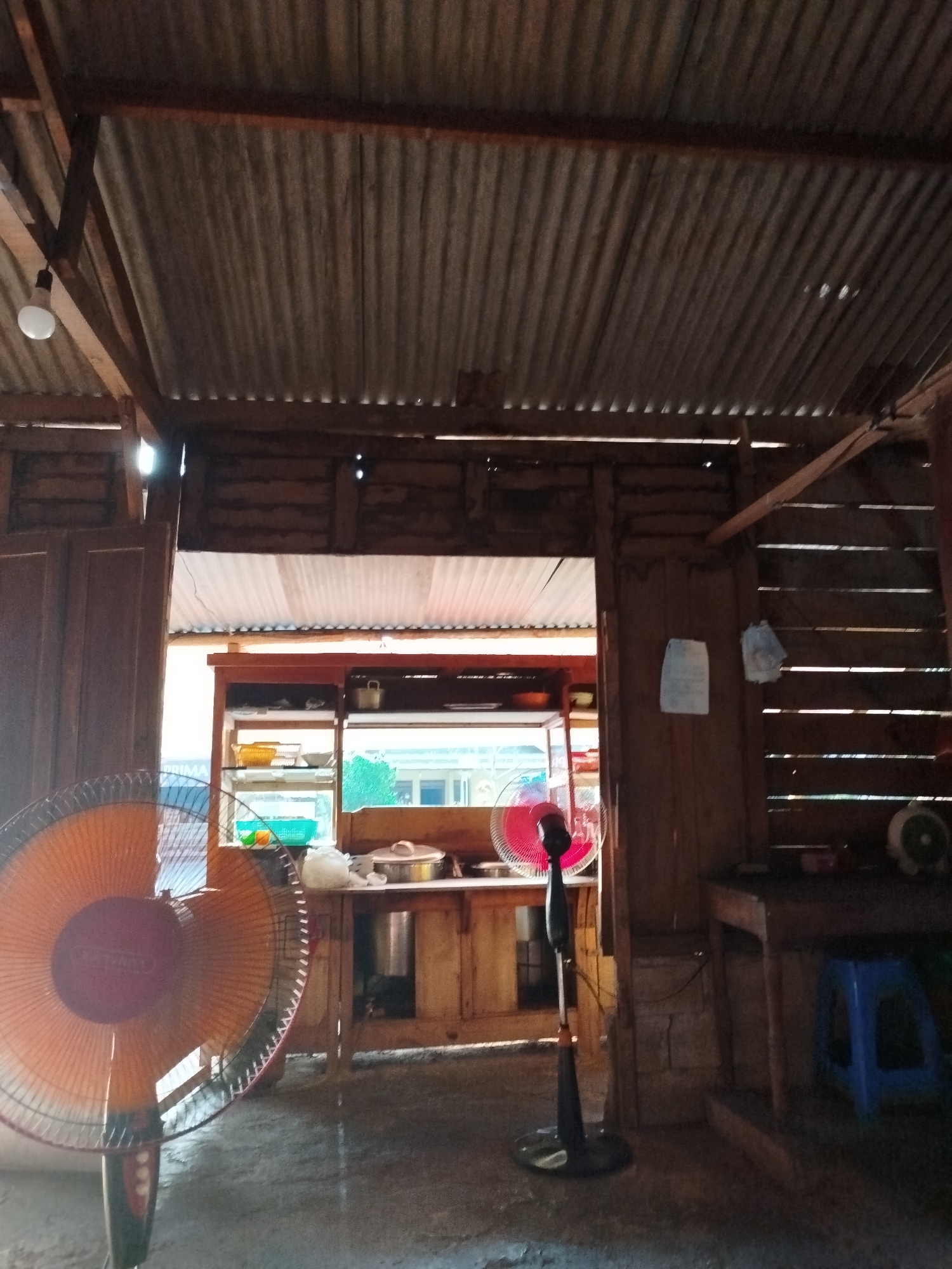 The image depicts the interior of a simple, rustic establishment, likely a food stall or small eatery, located in Pojok, Indonesia. The structure appears to be made primarily of wood and corrugated metal, suggesting a modest construction. In the foreground, a large, orange oscillating fan is prominently displayed on the left, angled towards the viewer. To the right of this fan, a smaller, black oscillating fan stands near a counter area.
Behind the fans, a counter is visible, stocked with various items. Shelves on the counter hold plates, bowls, and what appears to be a green plastic basket. A window behind the counter offers a glimpse of greenery and possibly other buildings outside. On the counter itself, there are some food items or ingredients, including a pot with a lid and some white objects that could be packaging or food preparation materials.
The walls of the establishment are constructed from horizontal wooden planks, and the ceiling is made of corrugated metal sheets, supported by wooden beams. A bare light bulb hangs from the ceiling on the left side. Two pieces of paper, possibly notices or menus, are attached to a wooden partition on the right wall. To the far right, a wooden table or counter is visible, with a small blue stool underneath. The floor appears to be concrete. The overall impression is one of a functional, utilitarian space designed for a small business in a warm climate.