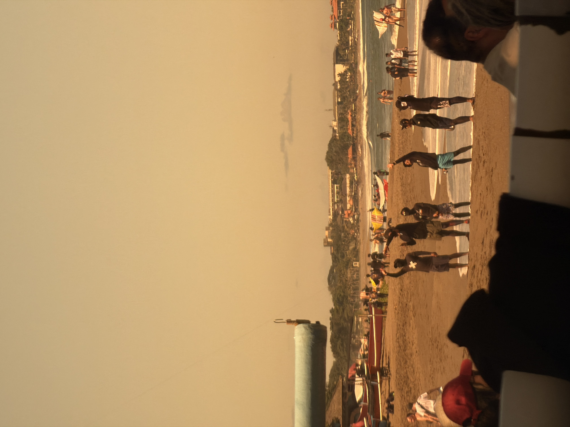 The image depicts a beach scene, possibly in the North Kuta region of Indonesia, given the context. The camera seems to be positioned slightly elevated and off-center, looking towards the shoreline.
In the foreground, there are partial views of objects and people that suggest the viewer is situated near a structure or seating area. There is a glimpse of a person's head and shoulder, a white object (possibly part of a table or structure), and some blurry dark shapes.
Moving towards the central part of the image, the beach is visible, where a group of individuals is standing. Some seem to be engaging in an activity that involves raising their arms, possibly in a form of exercise or routine.
The background features the coastline, where there are more people and the edge of the ocean meeting the sand. Further inland, there is a stretch of buildings and foliage, suggesting a developed coastal area. The sky is a uniform, light hue, possibly indicative of a clear day or the time of day.