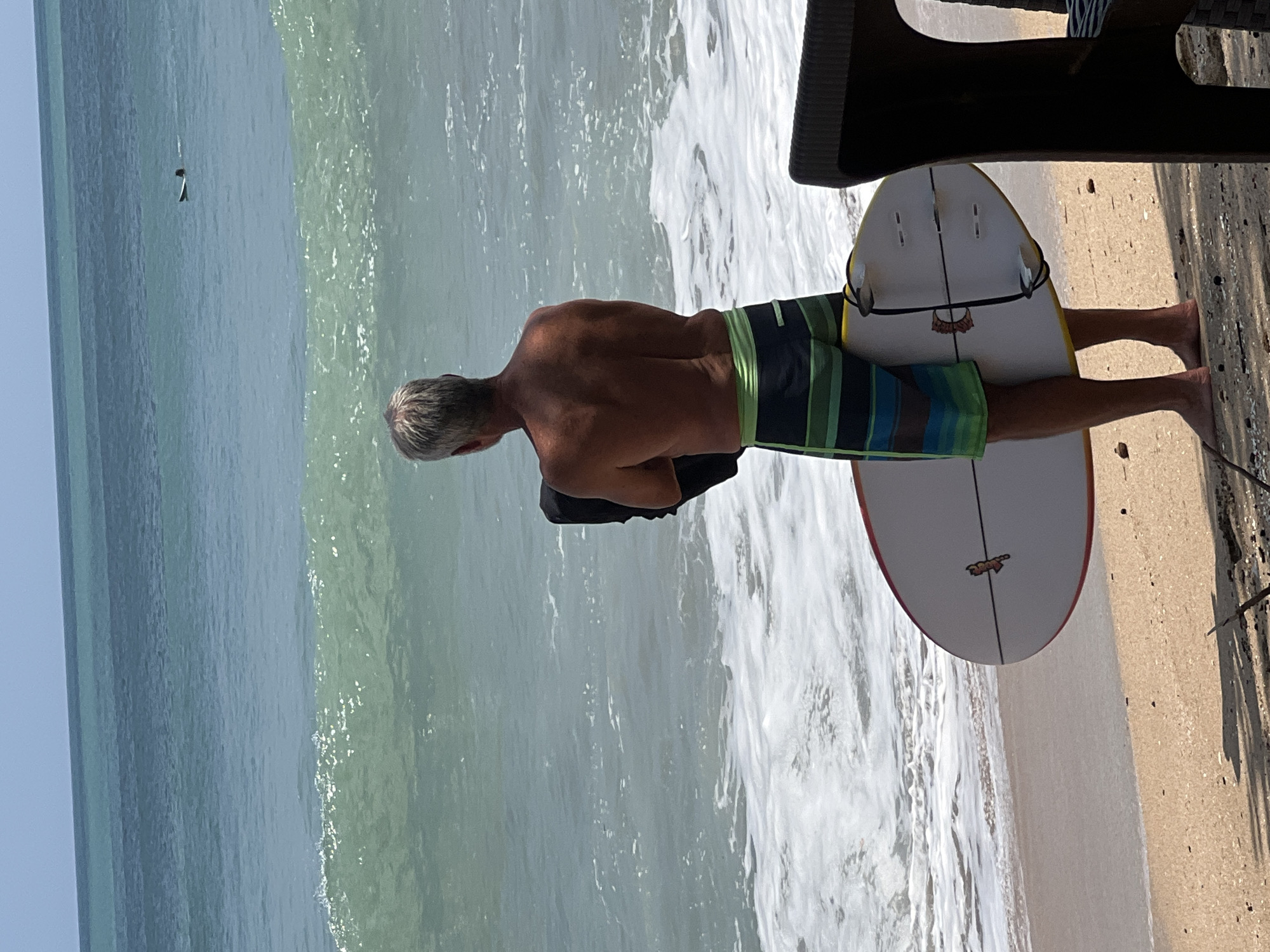 The image depicts a man on a beach in Kuta, Indonesia. He is standing with his back to the camera, holding a black bag in his left arm and a surfboard under his right arm. He is wearing colorful striped swim trunks and appears to be shirtless. The surfboard is white with a red and yellow trim and has three fins visible. The man is standing on a sandy beach with the ocean in the background. Waves are breaking on the shore, and the water is a gradient of blue and green. In the distance, another surfer is visible in the ocean. A portion of a dark-colored outdoor chair is visible in the foreground on the right side of the image. The sky is clear and blue. The scene captures a typical beach day in Kuta, known for its surfing spots.
