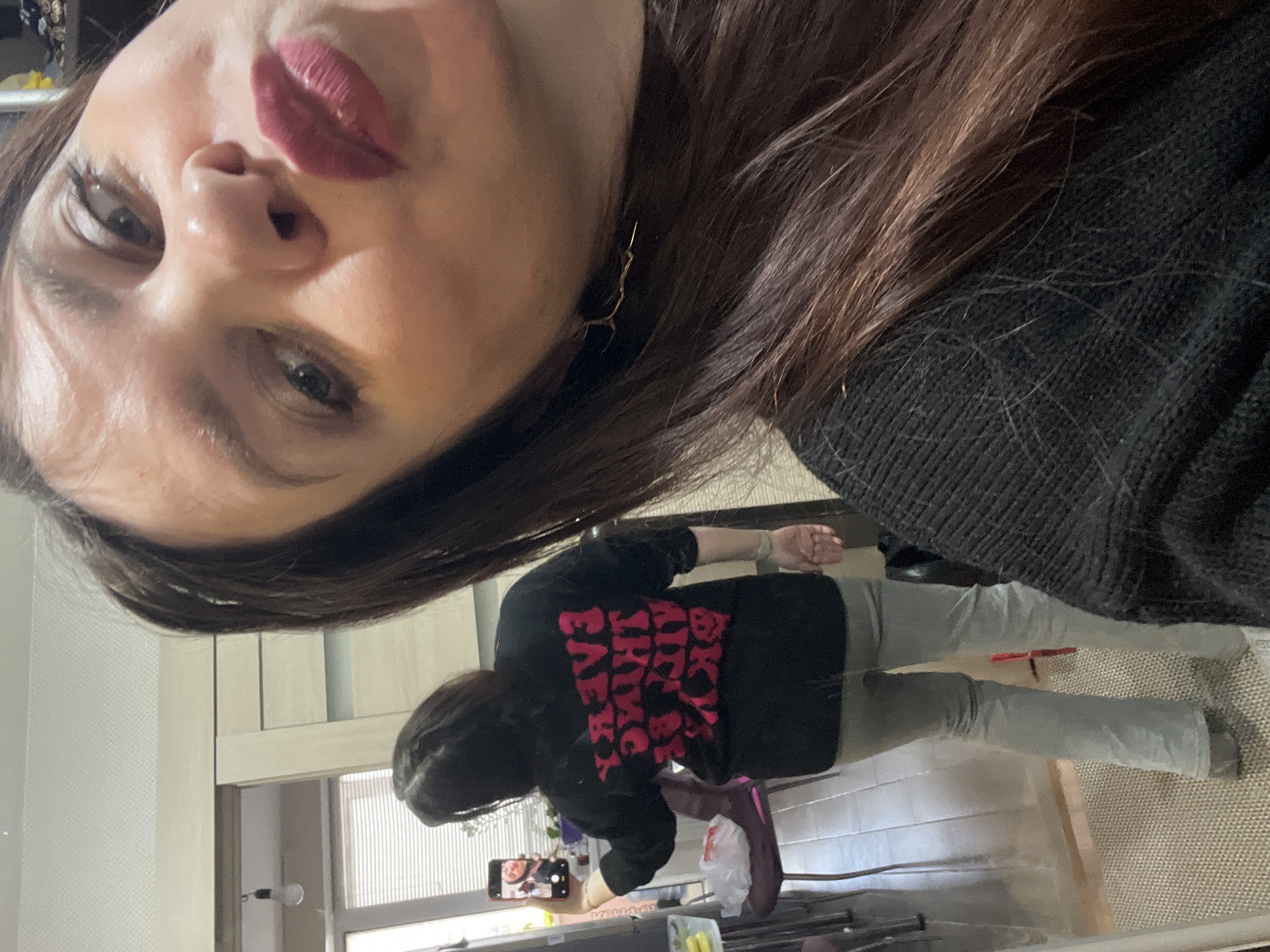 The image is a vertical selfie taken indoors, featuring a person in the foreground and their full-body reflection in a mirror in the background.
In the foreground, the upper half of a person's face is seen at a sharp upward angle. She has dark eyes, dark brown hair, and is wearing prominent dark red lipstick. A delicate, gold-colored earring, shaped like a small branch, hangs from her right ear. She is wearing a dark, possibly black, knit sweater or top. Her expression is neutral to slightly pensive.
In the reflection, the same person is visible standing. She has long, dark brown hair and is wearing a casual outfit consisting of a black short-sleeved t-shirt with bold pink text on the back, and light grey trousers or jeans. The text on the t-shirt reads "BLACK IS MY HAPPY COLOUR", with a skull and crossbones symbol between "HAPPY" and "COLOUR". She is holding a smartphone in her right hand, angled towards the mirror, indicating she is taking the photo. A blurred image of herself is visible on the phone screen. Her feet are partially obscured; one foot wears a dark shoe, and a purple bag or shoe is nearby.
The setting appears to be a domestic indoor space, possibly a bedroom or living area in Noviy, Russia. Elements in the background include a light-colored wall, a wooden doorframe, a window with vertical blinds, a visible lightbulb fixture, and a textured floor covering (possibly a rug or carpet). The lighting suggests daytime.