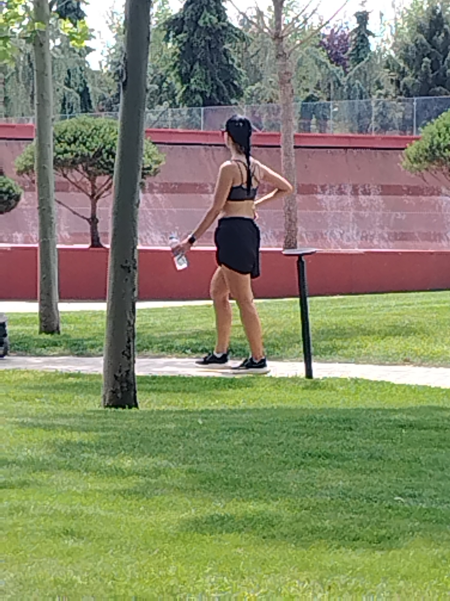 A woman is walking on a paved path in a park. She is wearing a black sports bra and black shorts, and her hair is pulled back in a ponytail. She is holding a clear water bottle in her right hand. The path is surrounded by green grass, and there are trees in the background. A dark, slender pole with a circular top is standing near the path. The background also shows a textured wall with a reddish hue and a fence. This scene is located in Krasnodar, Russia.
