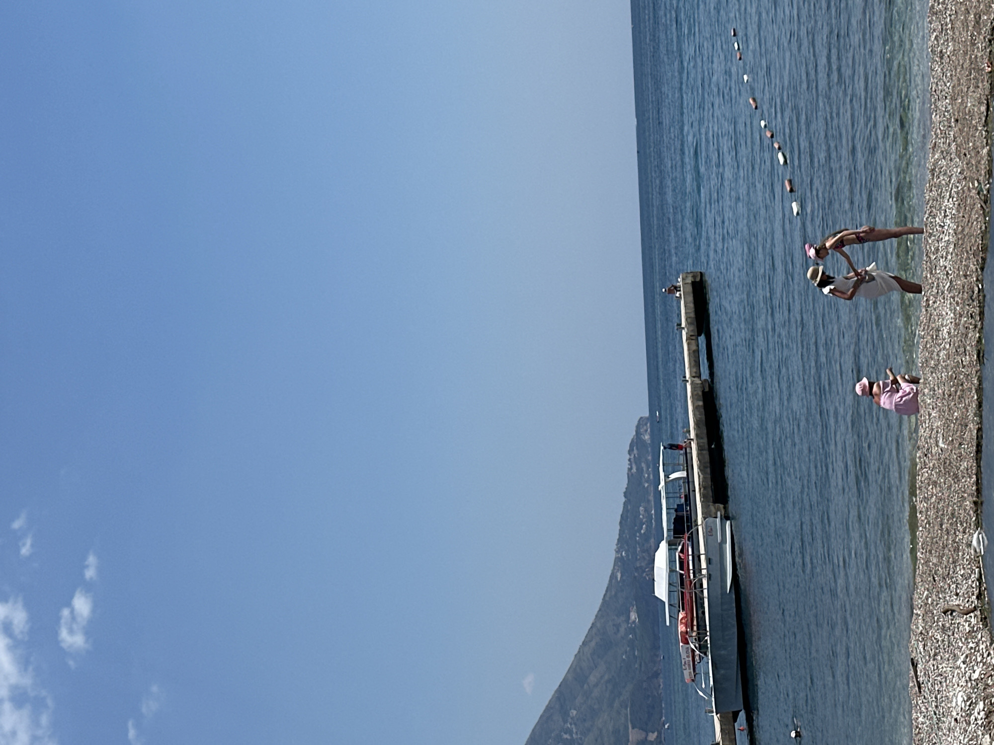 In Budva, Montenegro, on a sunny day, the scene is a beach with a clear blue sky and calm sea. In the foreground, a rocky shore leads to the water. A few people are present on the beach. To the left, a young person in a pink dress and hat is crouching near the water. To the right, two individuals, one in a white dress and hat and another in a bikini and pink hat, are standing and interacting. Further out in the sea, a pier extends from a landmass, with two boats docked and a few people visible on the pier and near the boats. A buoy line demarcates an area of the sea. The distant background features a verdant, mountainous coastline.