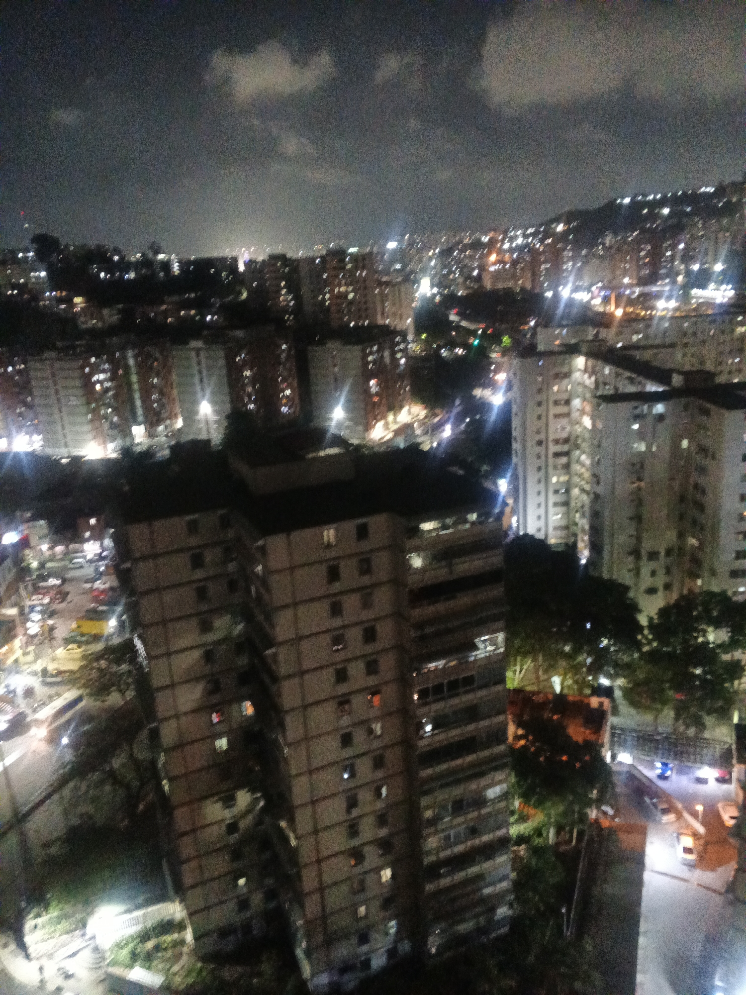 A nighttime view from an elevated position, possibly from a window or balcony, showcasing a dense urban cityscape. The scene is dominated by high-rise buildings, some of which are closer to the viewer and in sharper focus, while others fade into the background with blurred lights suggesting their distance. The buildings are illuminated by interior lights visible through windows. Streets are visible with moving car headlights creating streaks of light. Trees are present around some of the buildings, and beyond the city lies a low mountain range dotted with lights. The sky is dark, with patches of clouds illuminated from below.