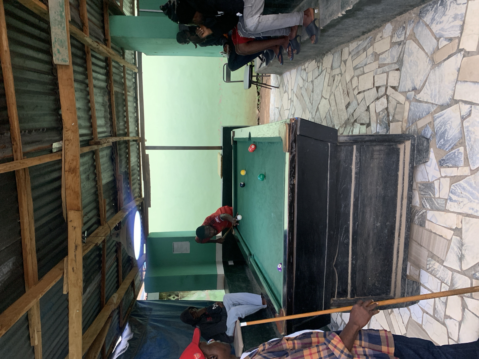 The scene shows a partially enclosed, open-air pool hall located in New Stadium, Nigeria. A pool table dominates the center, with a man in a red shirt aiming to strike the cue ball. Several other men are present; some are seated or leaning against a wall, while another holds a pool cue. The pool hall has a rudimentary structure with a corrugated iron roof supported by wooden beams. The surrounding walls are painted a light green. The floor is made of irregular stone tiles. A light fixture is visible attached to the corrugated iron roof.