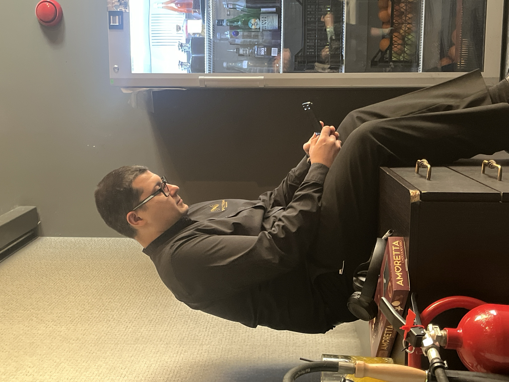 A man is sitting on a dark-colored trunk in what appears to be an office or break room. He is wearing a black button-down shirt, dark pants, and glasses. He is holding a small black object in his hands, possibly a phone or a remote control. Behind him is a refrigerator with bottles and snacks visible inside. Near the trunk is a red fire extinguisher, a box of "AMORETTA" chocolates, and a pair of headphones. The room has light-colored carpet and grey walls. The location is in the city of Sofia, Bulgaria.