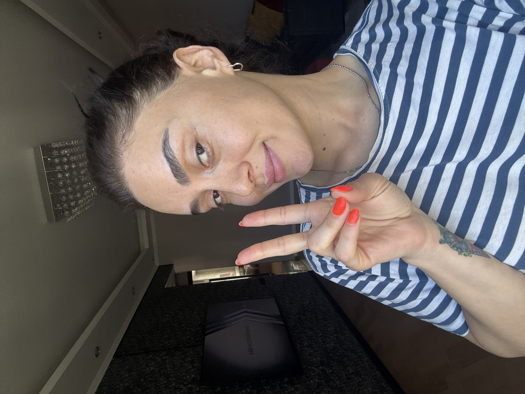 In this indoor, well-lit, eye-level medium shot, a woman is shown from the chest up, posing with her left hand raised in a peace sign, facing the camera. She has dark hair pulled back, a fair complexion, dark eyebrows, and appears to be wearing minimal makeup. Her nails are painted a vibrant orange. She is wearing a blue and white striped t-shirt and a silver chain necklace. A tattoo of a butterfly is visible on her left forearm. In the background, to the left, is a dark, textured wall with a framed graphic that reads "<<HАСТИЯ КИНО>>". To the right of the woman, a glimpse of a living room is visible, including furniture and a ceiling light fixture. The scene appears to be a residential setting, likely in Tbilisi, Georgia.
