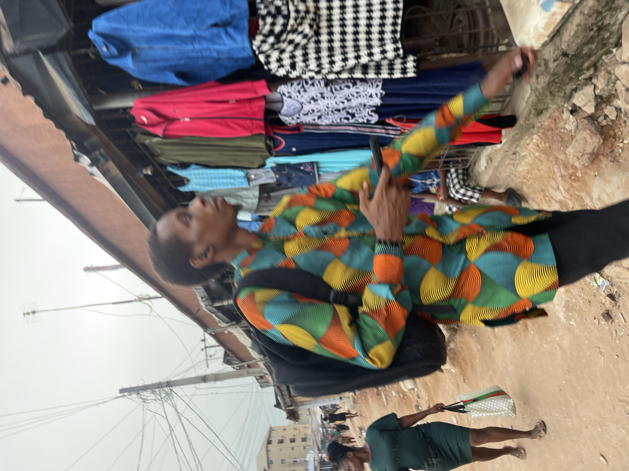 This image depicts a bustling outdoor market scene, likely in Onitsha, Nigeria. The primary subject is a person, possibly male, positioned on the left side of the frame, facing slightly away from the viewer. They are wearing a striking long-sleeved shirt adorned with a vibrant geometric pattern in shades of green, blue, yellow, and orange. A black backpack is on their shoulders. They hold a dark-colored smartphone up with their right hand, suggesting they are taking a photo or recording.
Behind this individual, racks of clothes are visible, indicating market stalls. Garments include a blue jacket, a red jacket, green fabric, light blue patterned fabric, a black and white checkered item, and white lace-patterned fabric.
In the mid-ground on the right, a woman walks away from the camera. She wears a dark green short dress and flip-flops, carrying a light-colored tote bag with red and green stripes. Several other indistinct figures can be seen further in the background, adding to the impression of a busy street.
The ground is unpaved, consisting of dirt and scattered rocks, characteristic of an informal market setting. Overhead, against an overcast sky, numerous utility poles are visible, crisscrossed with a dense network of electrical wires. The scene conveys a daytime atmosphere in a vibrant, active urban market. No visible text is discernible in the image.
