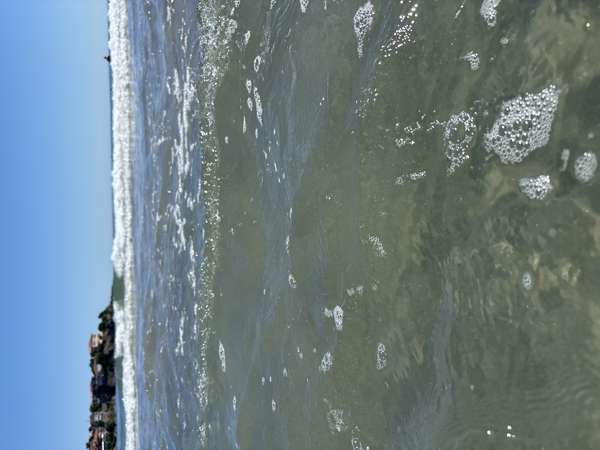 A close-up shot of the ocean water, with a blurry background showing waves, a coastline with buildings, and a clear blue sky. The water in the foreground is murky green and appears shallow, with some rocks visible on the seabed. Bubbles are scattered across the surface of the water. In the distance, a few indistinct figures can be seen in the ocean, possibly swimming or surfing.