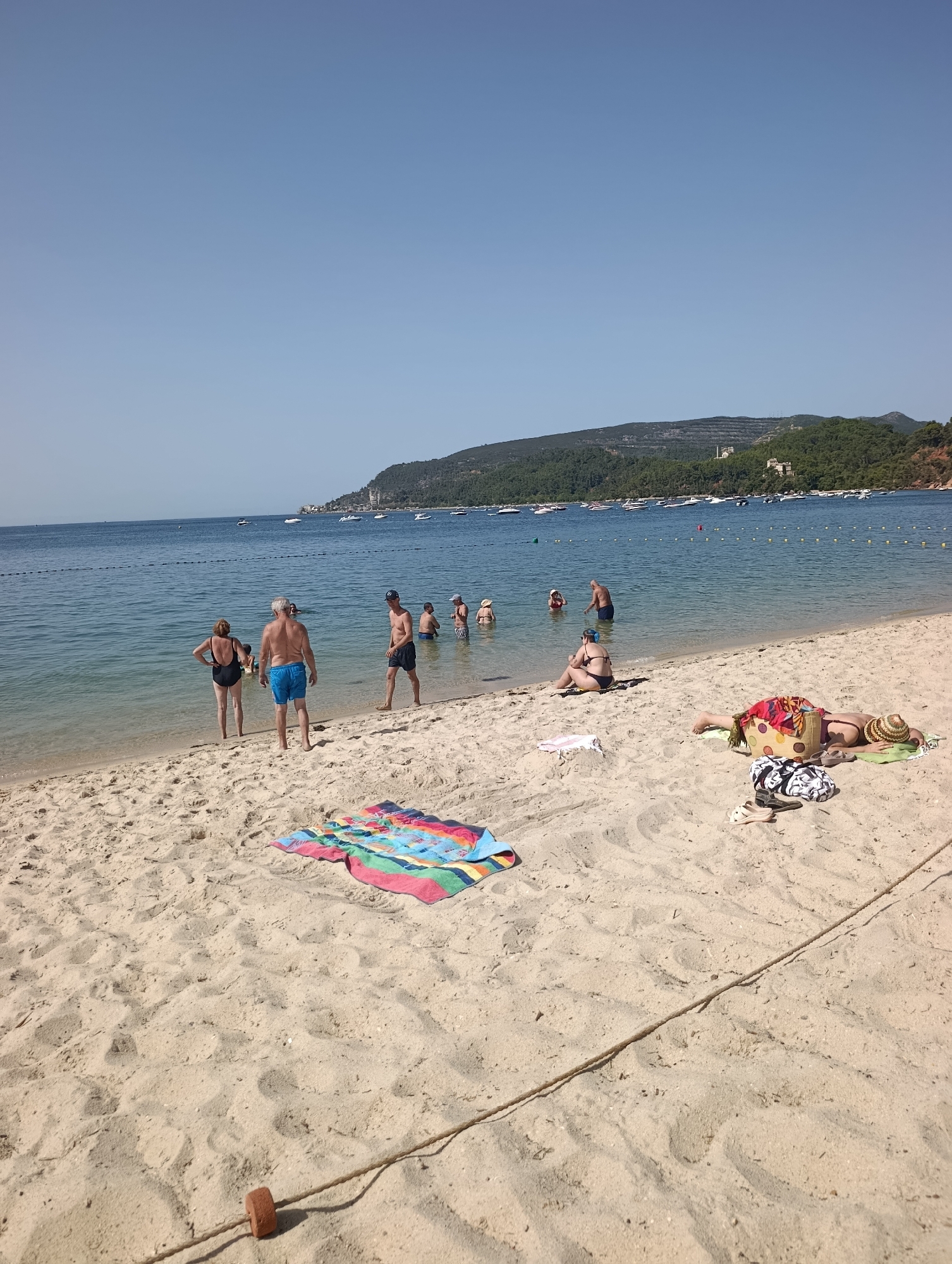 This image depicts a beach scene, likely in the city of Setúbal, Portugal, given the presence of the Arrábida mountain range in the background. The foreground is dominated by light-colored sand, with a brightly colored striped towel spread out on it. To the right, a person wearing a sun hat and a bikini is lying down on a towel, with a patterned bag and sandals nearby. Further towards the water, another person is sitting on the sand, also in beach attire.
In the shallow water, several individuals are visible. Two adults, a man in blue swim trunks and a woman in a black swimsuit, stand near the shore, facing away from the camera. Behind them, a group of people, including adults and what appear to be children, are wading or swimming in the sea. The water is clear and calm, with a few buoys marking a boundary in the distance.
The middle ground shows a calm sea dotted with numerous white boats anchored or moving slowly. The background features a lush, green, and hilly coastline, characteristic of the Arrábida Natural Park region near Setúbal. The sky is a clear, light blue. The overall impression is of a sunny, relaxed day at the beach.