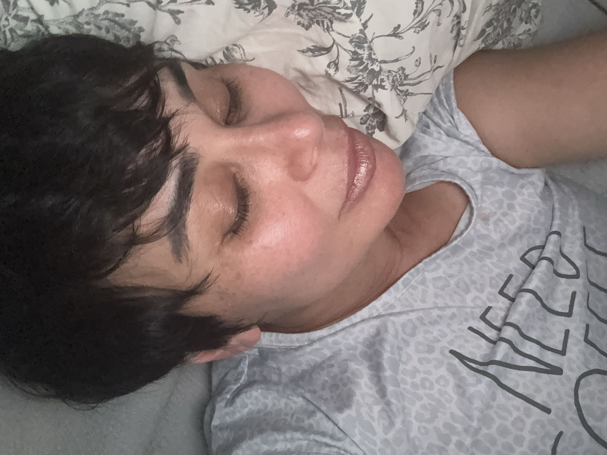 A close-up, overhead shot shows a woman lying on her right side on a bed, her eyes closed, and her head resting on a pillow. The woman has short, dark hair and is wearing a light gray t-shirt with a subtle leopard print. The t-shirt also has a dark gray graphic that reads "NEED COFFEE". The pillow beneath her head has a floral pattern in shades of gray and white. The background shows a portion of a white comforter. The scene appears to be indoors.