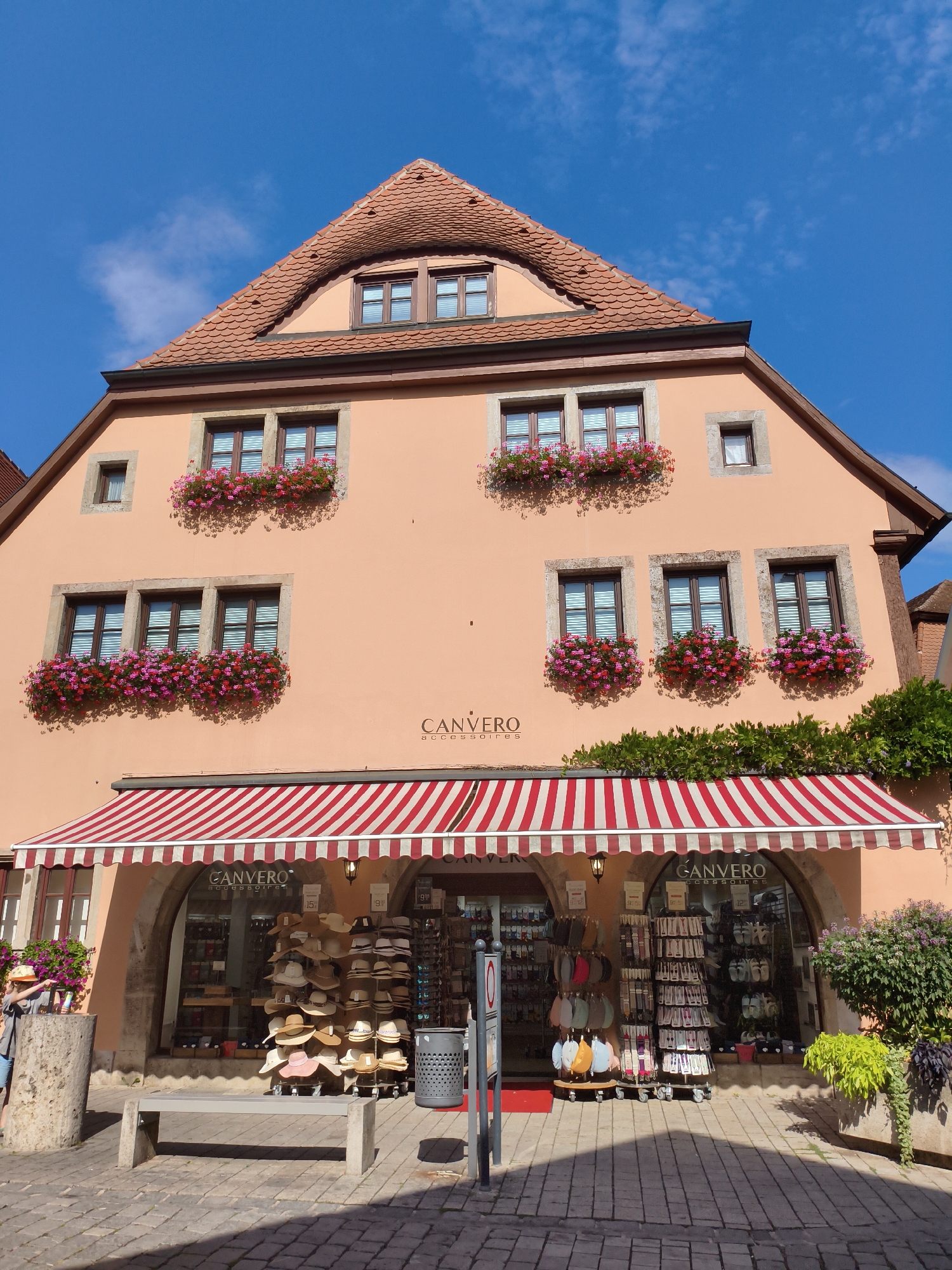The image shows the exterior of a shop called "CANVERO ACCESSOIRES" in Rothenburg ob der Tauber, Germany. The building is a multi-story structure with an orange facade and a red-tiled roof. The windows on the upper floors are adorned with flower boxes filled with pink and red flowers. A red and white striped awning covers the storefront, which displays a variety of hats and accessories for sale. A bench is placed in front of the shop, and a trash can is located to the right. The ground is paved with cobblestones, and the sky is a clear blue with a few scattered clouds. A person is visible on the left side of the image, near a stone pillar, looking at the flowers.
