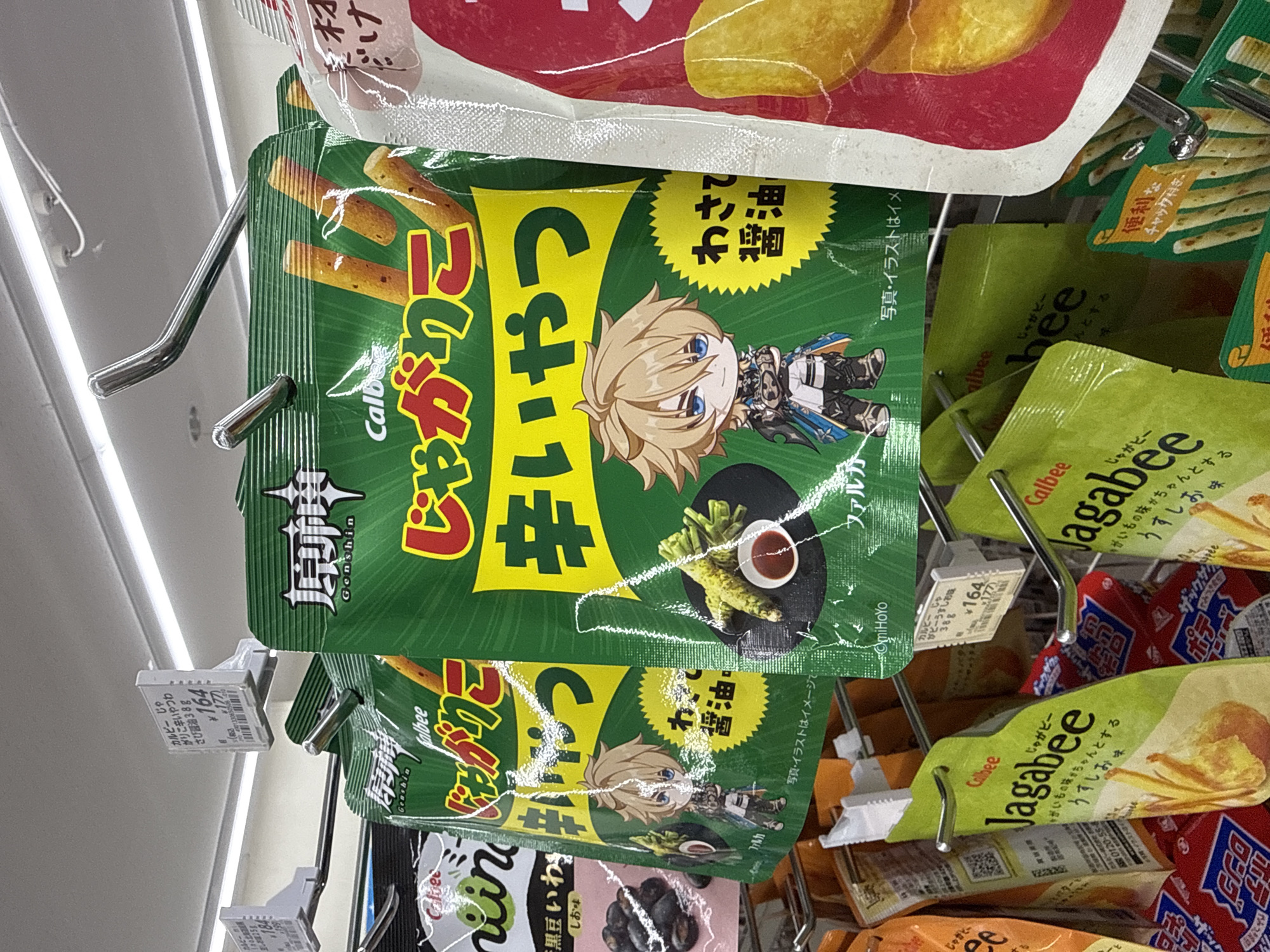 The image shows a close-up view of snack bags hanging on a retail display in a store. The primary focus is on a green snack bag with an anime-style illustration of a blond-haired character. The bag has Japanese text printed on it. Several other snack bags, mostly green and red, are visible in the background, also hanging on metal hooks. Some of these bags have Japanese text and brand logos. One bag is clearly labeled "Calbee Jagabee." A price tag with Japanese text and a price of "¥164" is attached to one of the bags. The scene appears to be an aisle in a convenience store or supermarket, likely in Japan, given the presence of Japanese text and the "Yachiyo, Japan" location context. The lighting suggests an indoor setting, possibly during daytime. The overall impression is a retail environment showcasing various snack products.