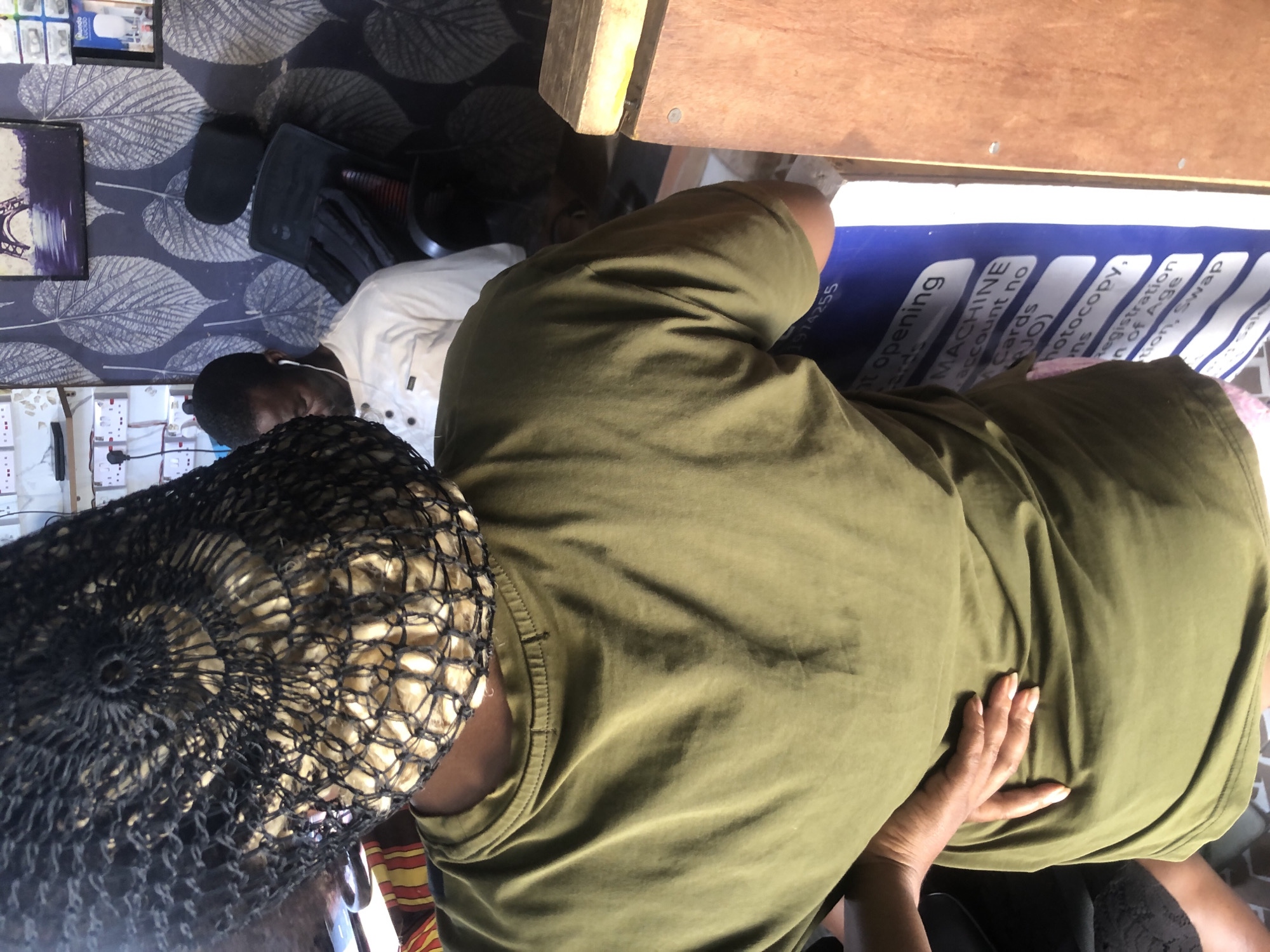 The image depicts an indoor scene, likely a small business establishment in Ado, Nigeria. In the foreground, a person wearing an olive green t-shirt and a black hair net is partially visible from the back, with their right hand resting on their hip. Behind this person, a man in a white collared shirt with earphones is seated. The background reveals a wall with a patterned wallpaper featuring large leaves, and shelves with electrical outlets and some items. A framed picture of the Eiffel Tower is also visible on the wall. In the lower right of the frame, a wooden counter or shelf is present. A blue sign with white text is partially visible, listing services such as "MACHNE", "account no", "Cards", "photocopy", and "Registration". The overall impression is of a bustling, everyday business environment.