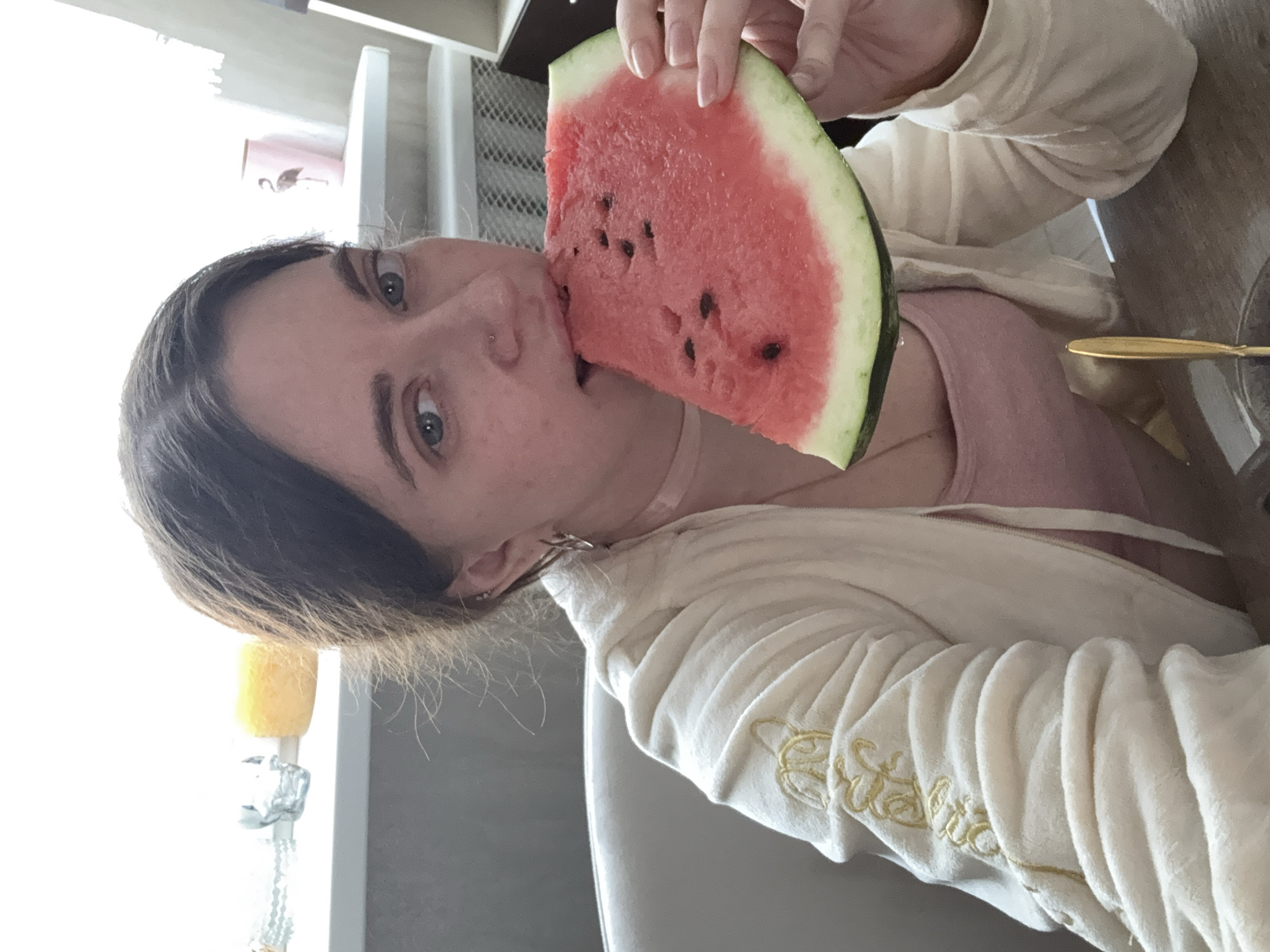 In an indoor setting, a person with dark hair and blue eyes is captured in a close-up shot, biting into a slice of watermelon. They are wearing a cream-colored hooded jacket with gold embroidered detailing on the sleeve, and a pink tank top underneath. The person has a nose ring and earrings. The background shows a shelf with decorative items, including a candle and a small figurine. A table with a gold utensil is in the foreground. The lighting suggests an indoor environment during the day.