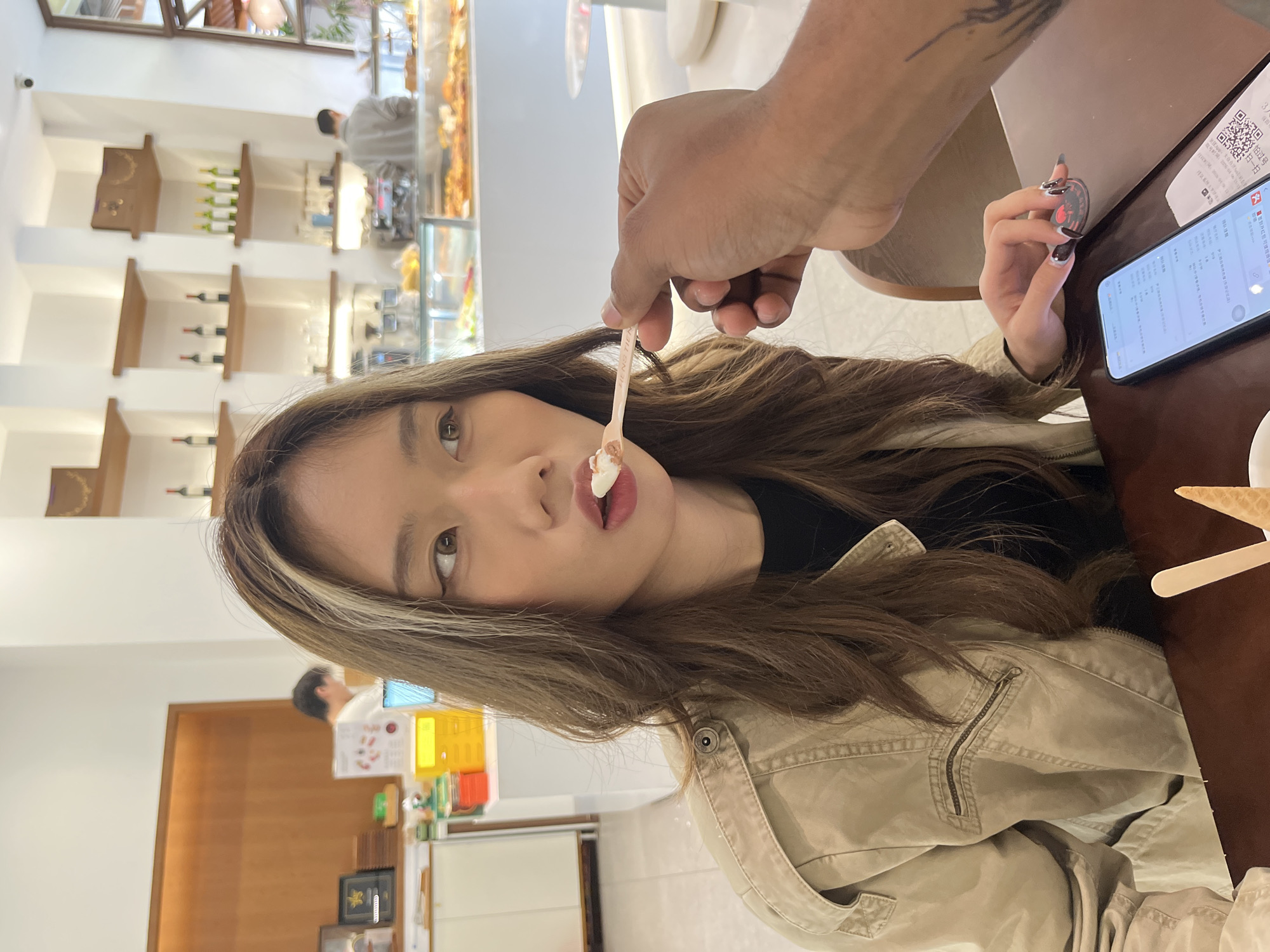 A woman with brown hair and a tan jacket is being fed ice cream by a dark-skinned hand in what appears to be a cafe. The woman is looking directly at the camera with ice cream on her lips. A smartphone is visible on the table beside her, along with a coffee cup with a spoon. Shelves of wine bottles and baked goods suggest a cafe or bakery setting. The location is indicated to be Chenghua District, China.
