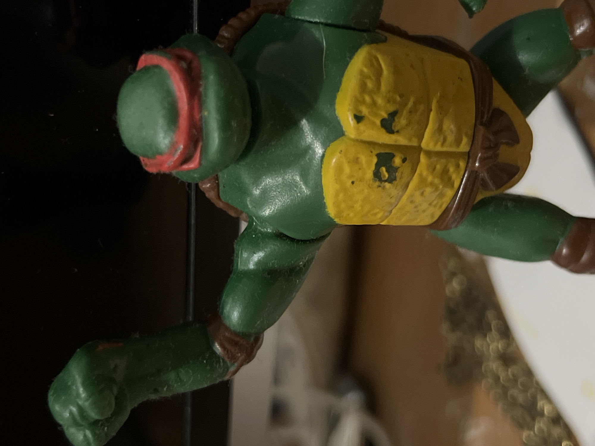 A close-up, vertically oriented photograph depicts a green Teenage Mutant Ninja Turtle action figure, identifiable as Raphael by its red bandana. The figure's body is a solid green, contrasting with a worn yellow plastron (belly shell) that exhibits several chips and scuff marks, revealing the green plastic underneath. Brown details, representing parts of the shell and limb straps, are visible around the shoulders, hips, and torso. Small dust particles and fibers are noticeable on the figure's surface, particularly on the head and arms, suggesting age or handling. The figure's head, with its red eye-mask, is in the upper left, while a clenched fist is visible on the lower left arm. The background is split between a dark, possibly reflective surface on the left and a blurred, lighter area with bokeh on the right. No specific activity is occurring, and there is no visible text. The lighting emphasizes the texture and aged condition of the action figure.