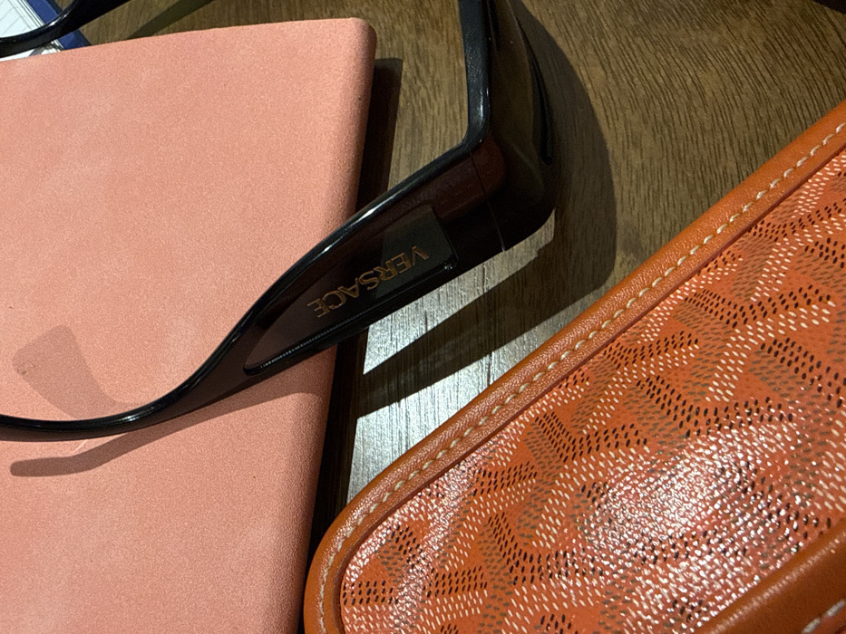A close-up shot captures a stylish tableau. Black Versace sunglasses rest atop a salmon-colored book or notebook. An orange Goyard wallet or pouch, edged with meticulous stitching, fills the lower right corner. The arrangement sits on a textured, likely wooden, surface. The composition highlights luxury brands and attention to detail.