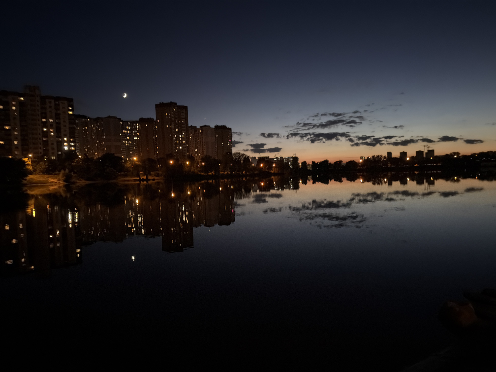 The image depicts a twilight scene over a body of water, likely a lake or river, with the skyline of Kyiv, Ukraine, in the background. The sky is a deep indigo fading to a pale orange and yellow near the horizon, indicating the last moments of daylight or the first moments before sunrise. A sliver of a crescent moon is visible in the upper left quadrant of the sky.
Along the left side of the image, several tall, multi-story apartment buildings are illuminated, with many windows showing interior lights. These buildings are characteristic of Soviet-era residential architecture common in Kyiv. The lights from the buildings are reflected in the dark, still water, creating shimmering patterns.
Further to the right, along the horizon, more buildings and structures, some appearing to be under construction with cranes, are silhouetted against the lighter sky. Scattered lights are visible within these structures as well. Trees and other vegetation line the shore in front of the buildings, also silhouetted.
The water's surface is calm, acting as a mirror for the lights and structures above, though some ripples and distortions are present, particularly in the reflections of the clouds. The overall mood is peaceful and serene, capturing the quiet transition from day to night in an urban setting. There are no people or overt actions visible in the image. The scene is consistent with an urban landscape found within Kyiv, Ukraine, perhaps a residential area adjacent to a recreational body of water.